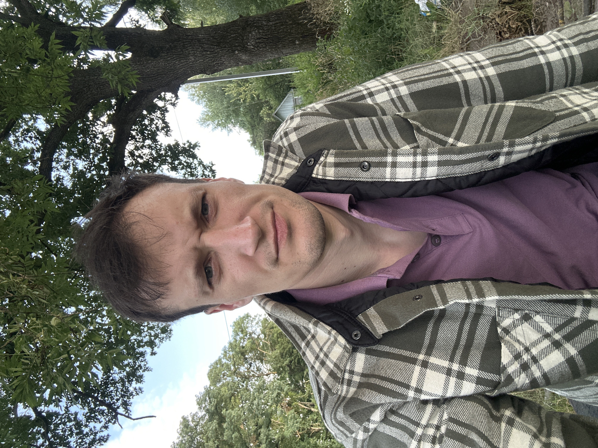 In Makiv, Ukraine, a young man with short, dark hair is pictured from the chest up, looking directly at the camera with a serious expression. He is wearing a purple polo shirt under a green and white plaid jacket with a black quilted lining. The background consists of lush green trees and foliage under a blue sky with scattered clouds, suggesting an outdoor setting. A large tree trunk is visible to the right of the man, and a rustic, light-colored building is partially visible in the background to the right. The overall lighting indicates it is daytime.