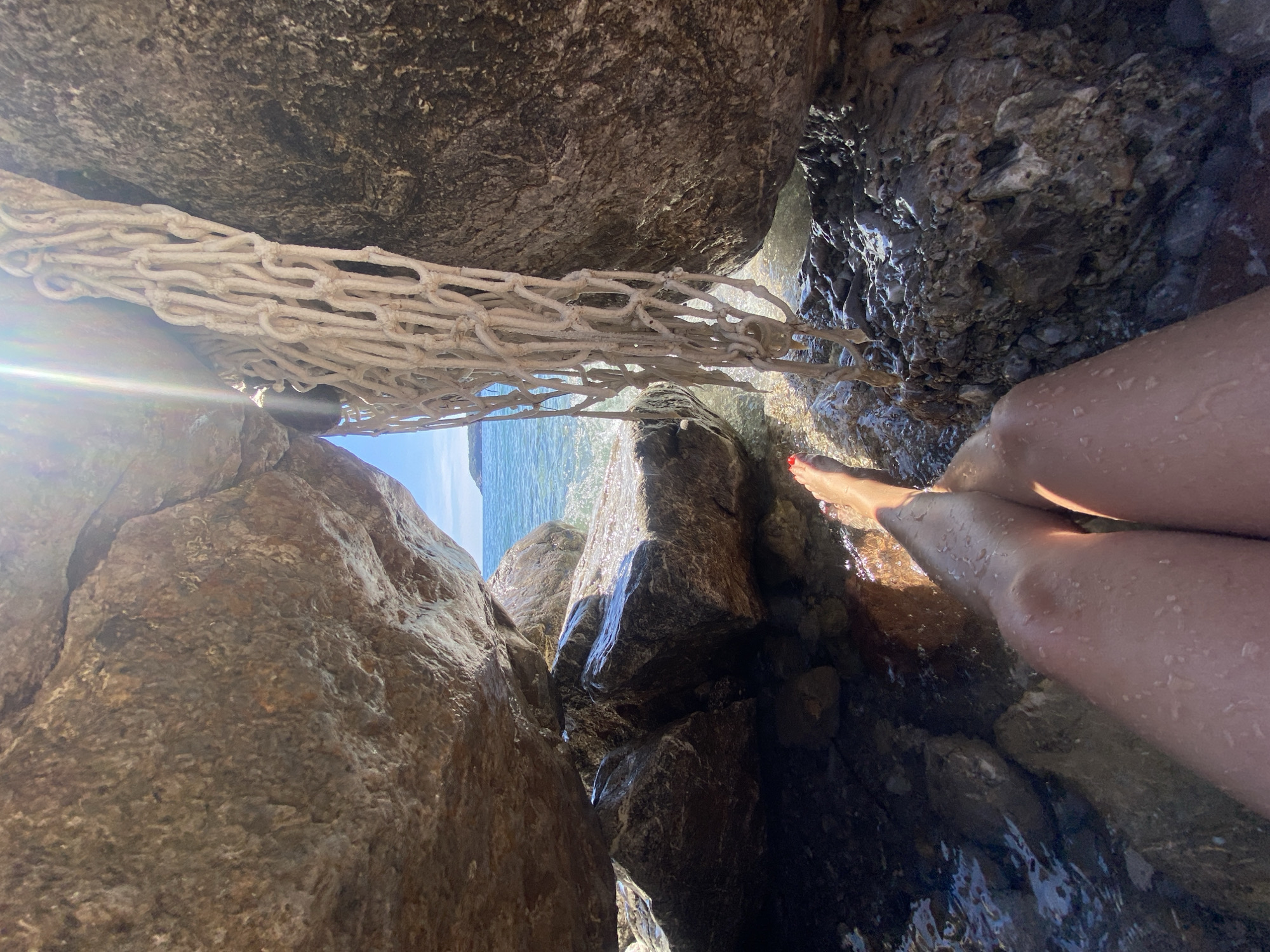 The image depicts a view from inside a narrow rock crevice at a beach. The perspective is upwards, looking out towards the sea through the gap between the rocks. The rocks are large and textured, primarily brown and gray. A white rope net is suspended across the opening. To the right side, a pair of legs, likely a woman's due to the presence of red nail polish on a toe, are visible near the water's edge. The water is clear and slightly illuminated by sunlight. The sea is visible in the distance, with a sliver of blue sky above.
There is no way to tell from the image whether or not it is in Prijevor, Montenegro. The description does not require that I know this information.