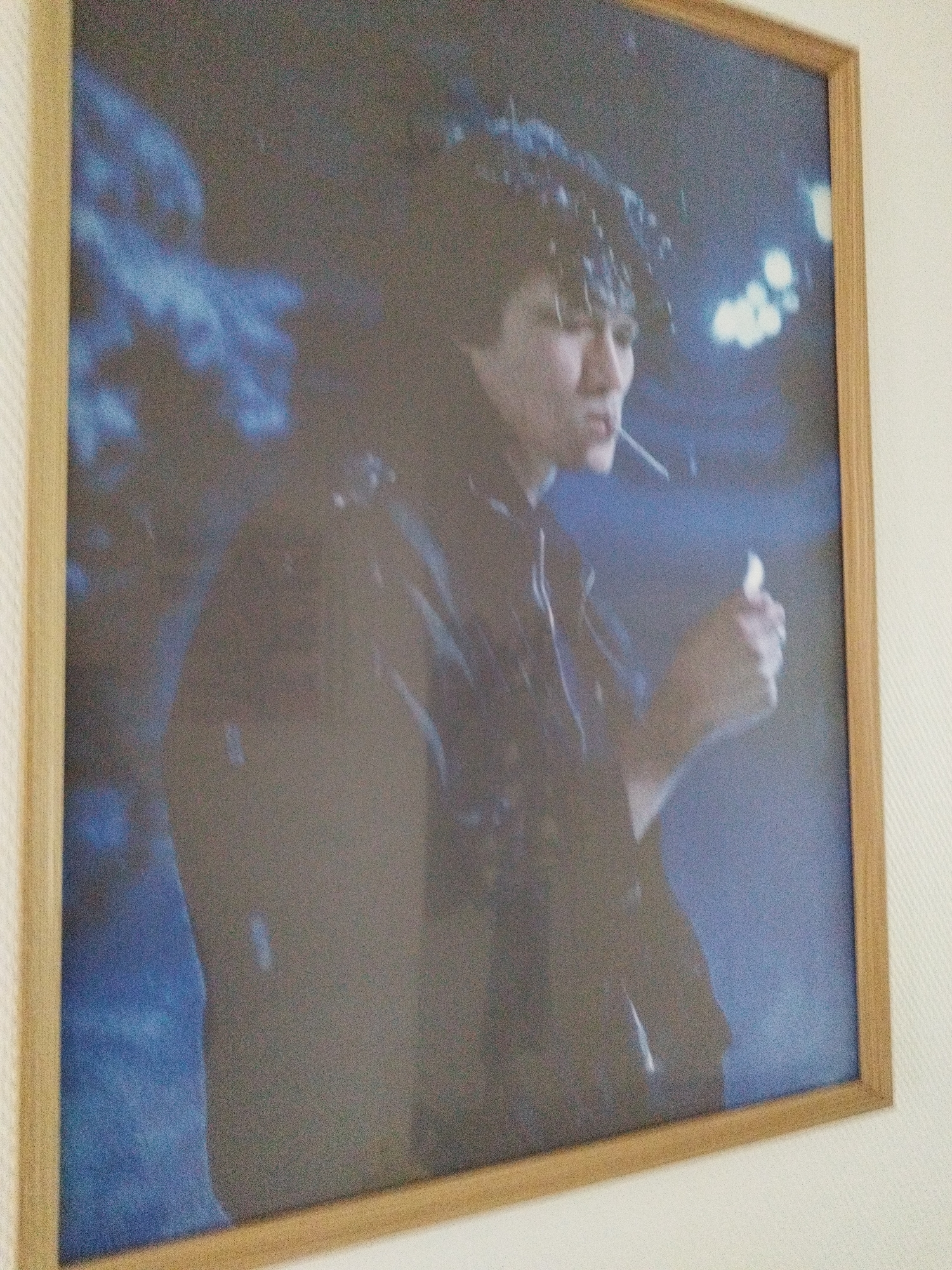 The image shows a framed portrait of a young person with dark, curly hair, in profile, smoking a cigarette. The background is dark blue, with blurry lights that suggest an outdoor, possibly nighttime, setting. The person is wearing dark clothing and appears to be looking away from the camera. The overall tone of the image is somewhat melancholic. The location is not discernible from the image itself.