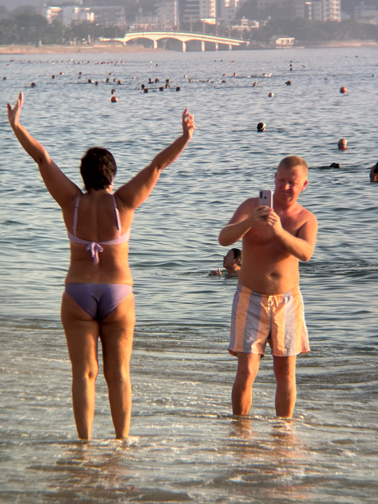 Two individuals are in shallow ocean water on a beach. The person on the left, viewed from behind, is a woman wearing a light purple bikini. She has her arms raised above her head. The person on the right, viewed from the side, is a man wearing striped swim trunks. He is holding a smartphone and appears to be taking a picture. In the background, other people are visible in the water, and a city skyline with buildings and a bridge can be seen in the distance. The water is calm with gentle waves. The scene is likely a public beach in Nha Trang, Vietnam, given the presence of the city skyline and the tropical setting.