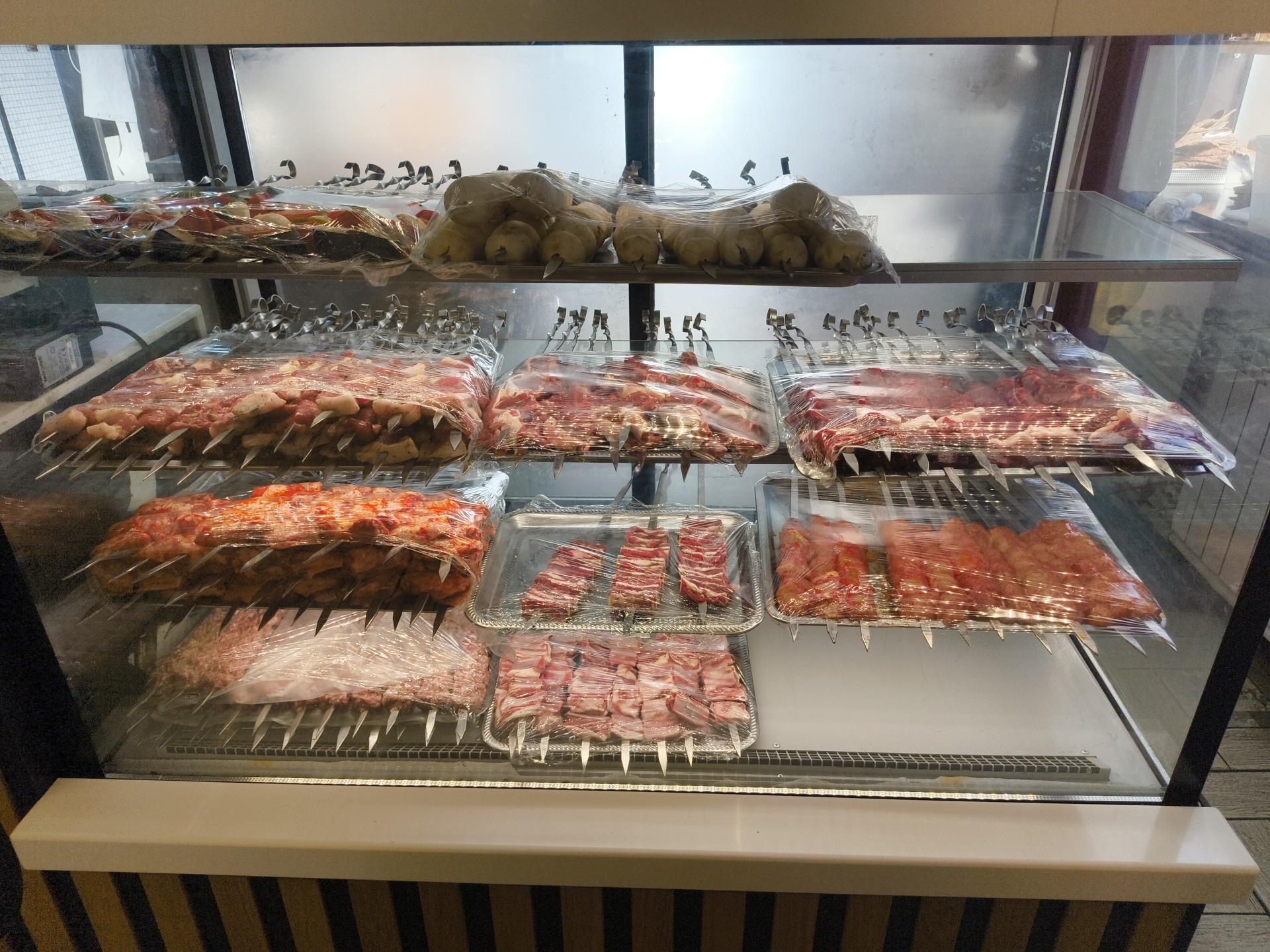 The image shows a refrigerated display case filled with various types of meat skewers, likely at a butcher shop or food market. The meat is raw and packaged in plastic wrap, with metal skewers inserted through it. There are several rows of skewers, with different cuts of meat visible, including what appears to be lamb chops, cubed meat, and possibly ground meat. The display case is clean and well-lit. The background is blurred but suggests an indoor setting with glass panels. There are no people visible in the image. The overall impression is one of a well-stocked food service establishment. This scene is not specific to Moscow, Russia and could be found in many similar establishments globally.