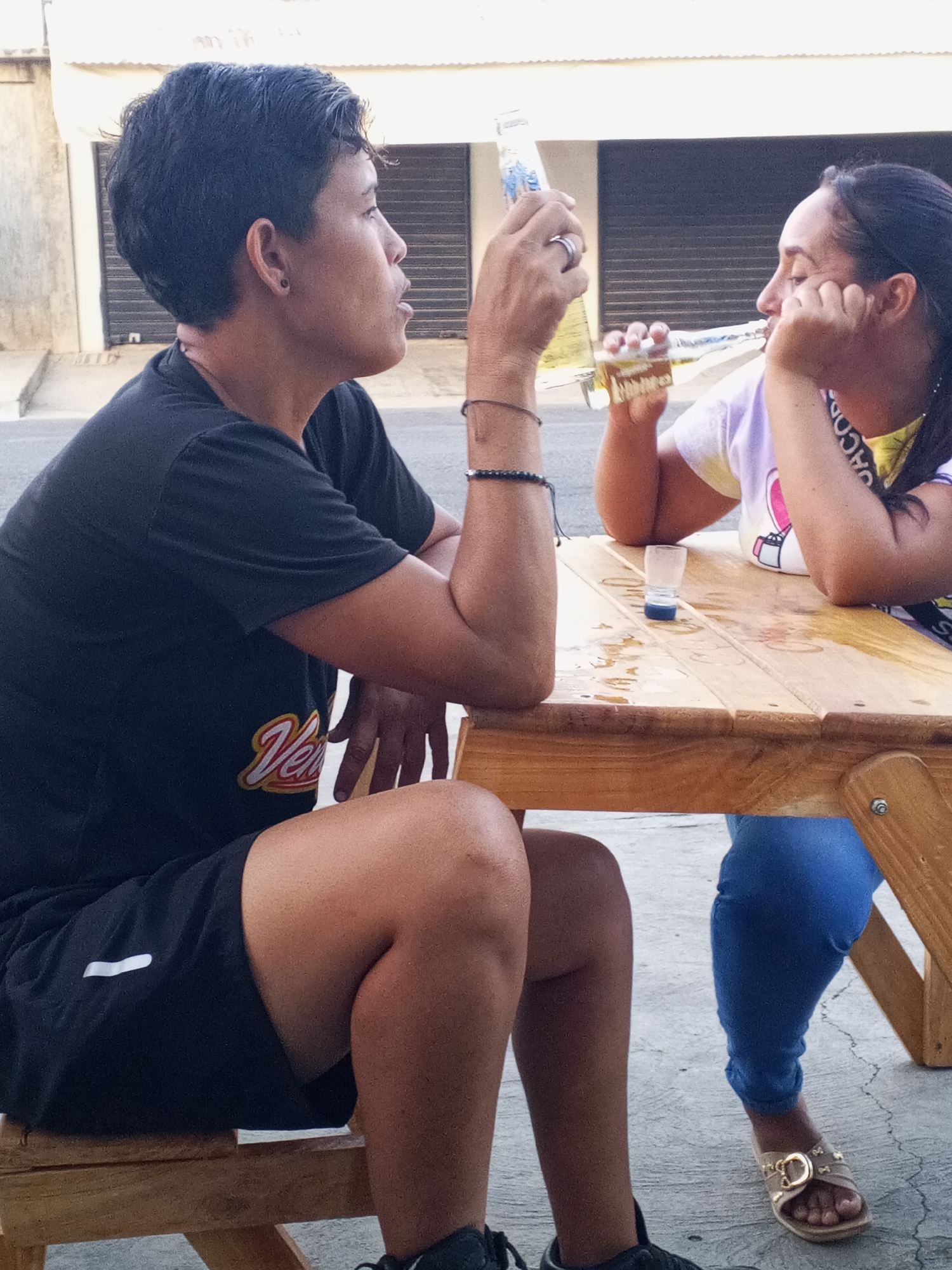Two individuals are seated at a light-colored wooden table, seemingly outdoors, in an urban environment. The person on the left has dark hair and is wearing a black t-shirt and black shorts. They are seated on a wooden stool and holding a bottle. The person on the right has dark hair pulled back and is wearing a light-colored t-shirt and blue pants. They are resting their head in their hand, also holding a bottle. There is a shot glass on the table between them. The background shows a building with closed roller doors. The location appears to be a street or public space in an unknown city.