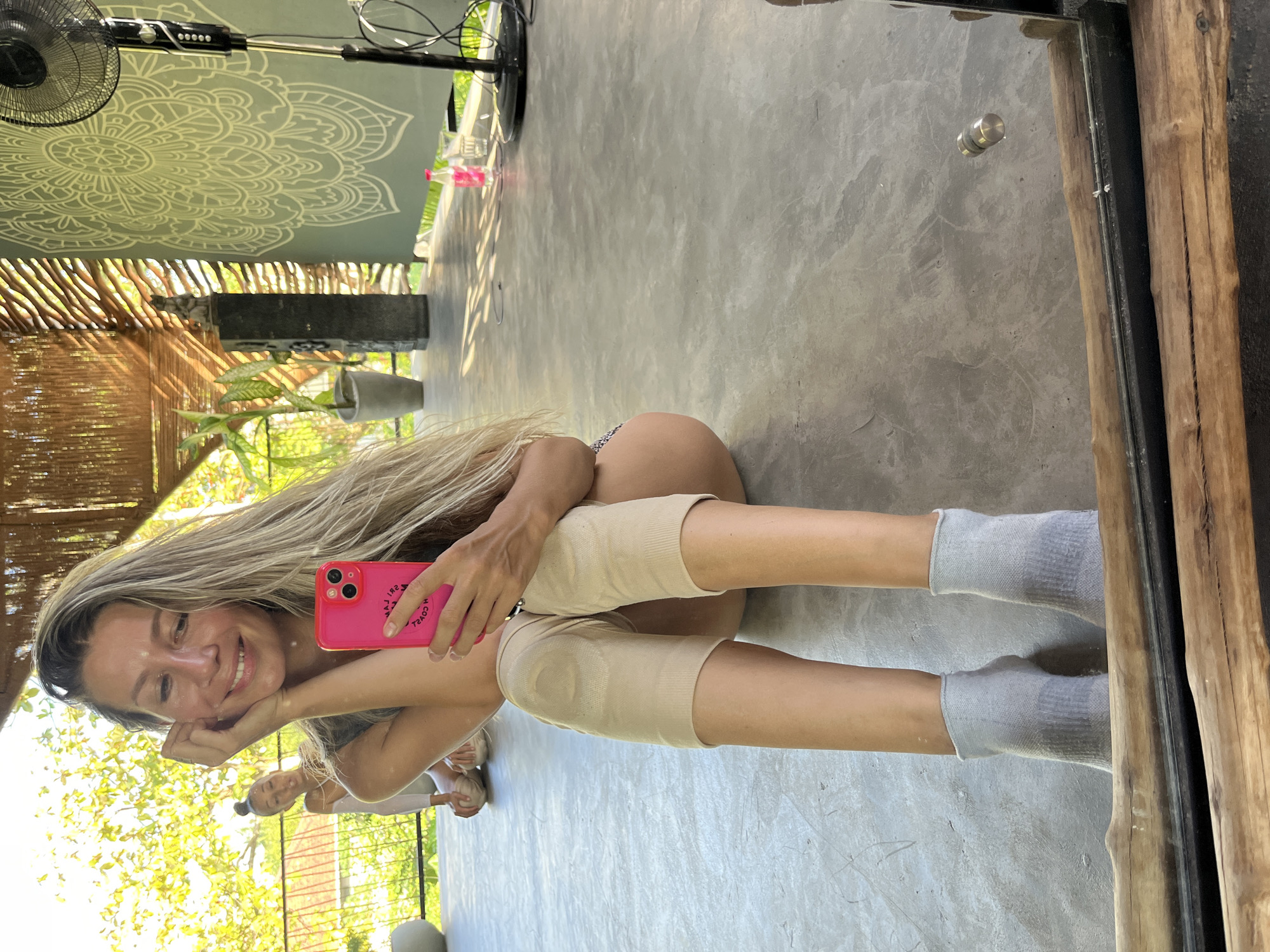 The image shows a woman sitting on a concrete floor, taking a selfie with a pink phone. She has long blonde hair and is smiling. She is wearing a gray tank top, beige knee sleeves, and gray socks. Another woman is visible in the background, also sitting on the floor. The setting appears to be an open-air yoga studio with a thatched roof and bamboo walls. A large mandala-like design is painted on a wall in the background, and a standing fan is also visible. The scene is brightly lit by natural sunlight. The location is identified as Weligama, Sri Lanka, likely a yoga studio or resort area.