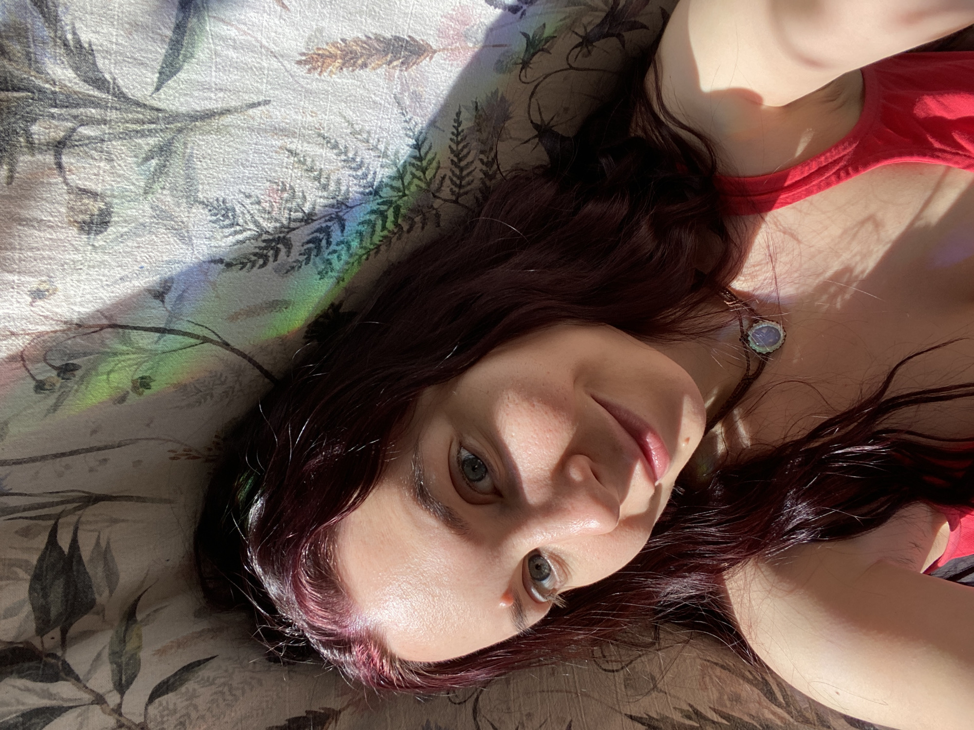 A fair-skinned woman with auburn-toned hair and light eyes lies on a floral-patterned surface, likely fabric, with a rainbow spectrum of light projected across it. She is wearing a red top and a necklace with a pendant. The location is the city of Chaikovsky, Russia. The woman is looking directly at the camera with a slight smile. The lighting is a mix of direct sunlight and shadows.