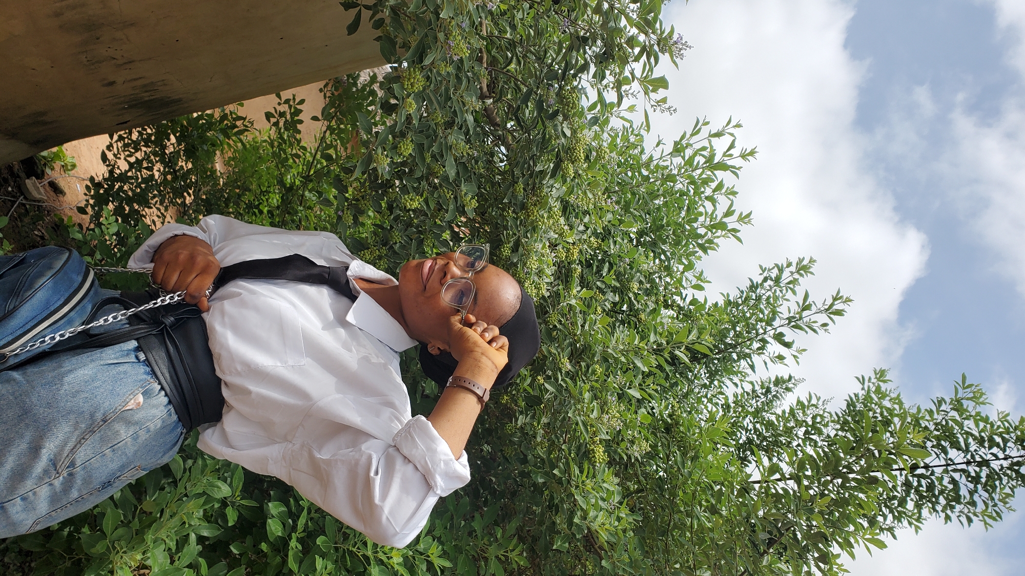 A young Black woman, wearing a white collared shirt, a black tie, clear-rimmed glasses, and a black headscarf, stands outdoors with a lush green bush behind her and a bright sky above. Her right hand is raised to her forehead as she looks away to her right, with a slight smile. She is wearing a brown watch on her left wrist. She has a black belt tied around her waist and a denim skirt. A blue and silver bag is slung over her shoulder by a silver chain. The background consists of dense green foliage and a cloudy sky.