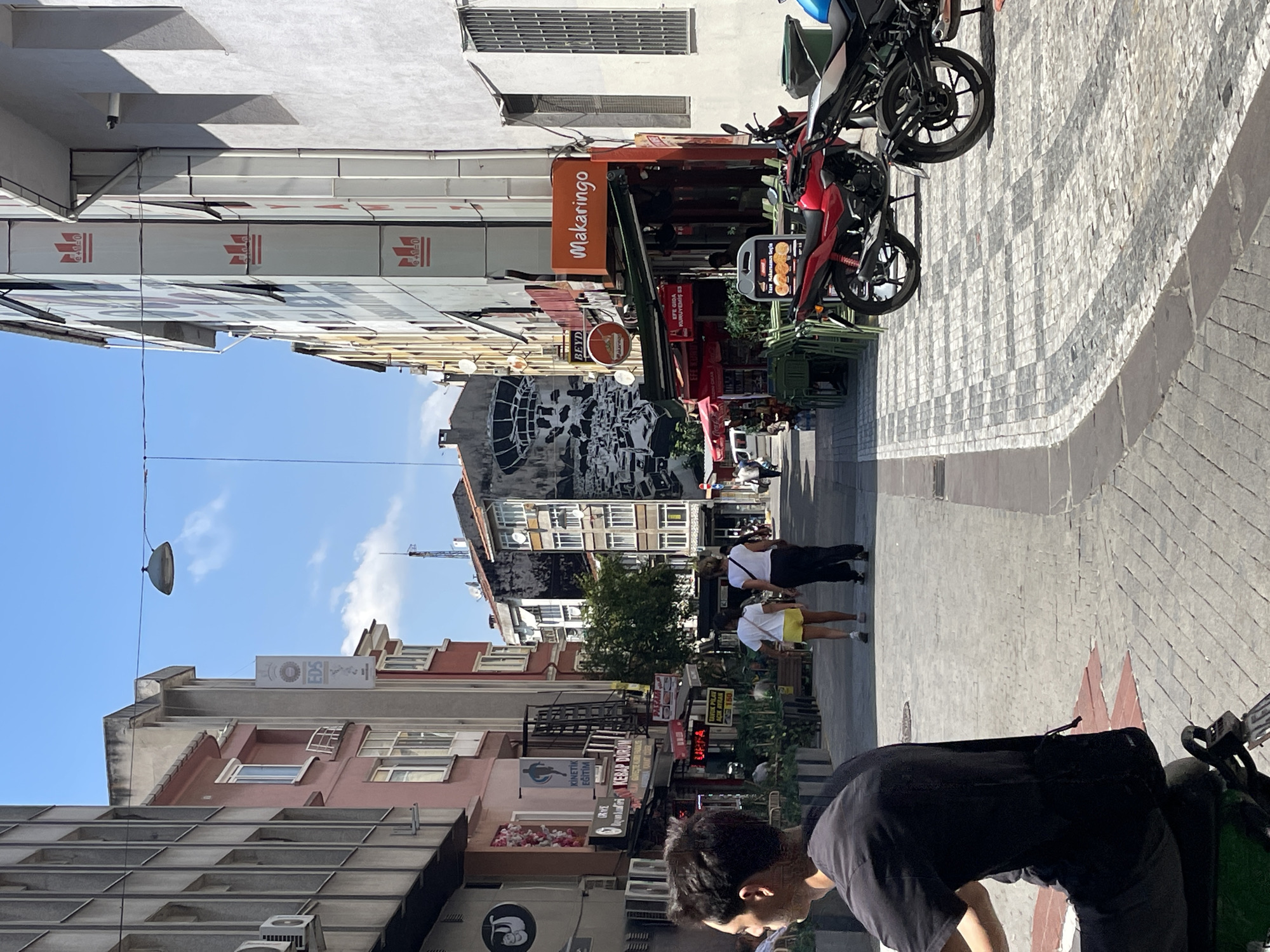 The image captures a street scene in Kadıköy, Turkey. In the foreground, a person is partially visible from behind, seated and facing away from the camera. The street is paved with cobblestones, with a checkered pattern of light and dark gray stones. On the right side of the street, several motorcycles are parked. A red motorcycle is prominent, followed by others. Shops line both sides of the street. On the right, "Makarino" is visible on an orange sign above a shop entrance, with tables and chairs set up outside. Further down the street on the right, a large mural depicting a futuristic city or stadium is visible on the side of a building. On the left side, a shop with a sloth logo is visible, along with signs for "Kinetic Egitim" and "Kebap Durum." Two individuals, a woman in a white shirt and yellow shorts and another person in a white shirt and black pants, are walking away from the camera down the street. The sky above is blue with some white clouds.