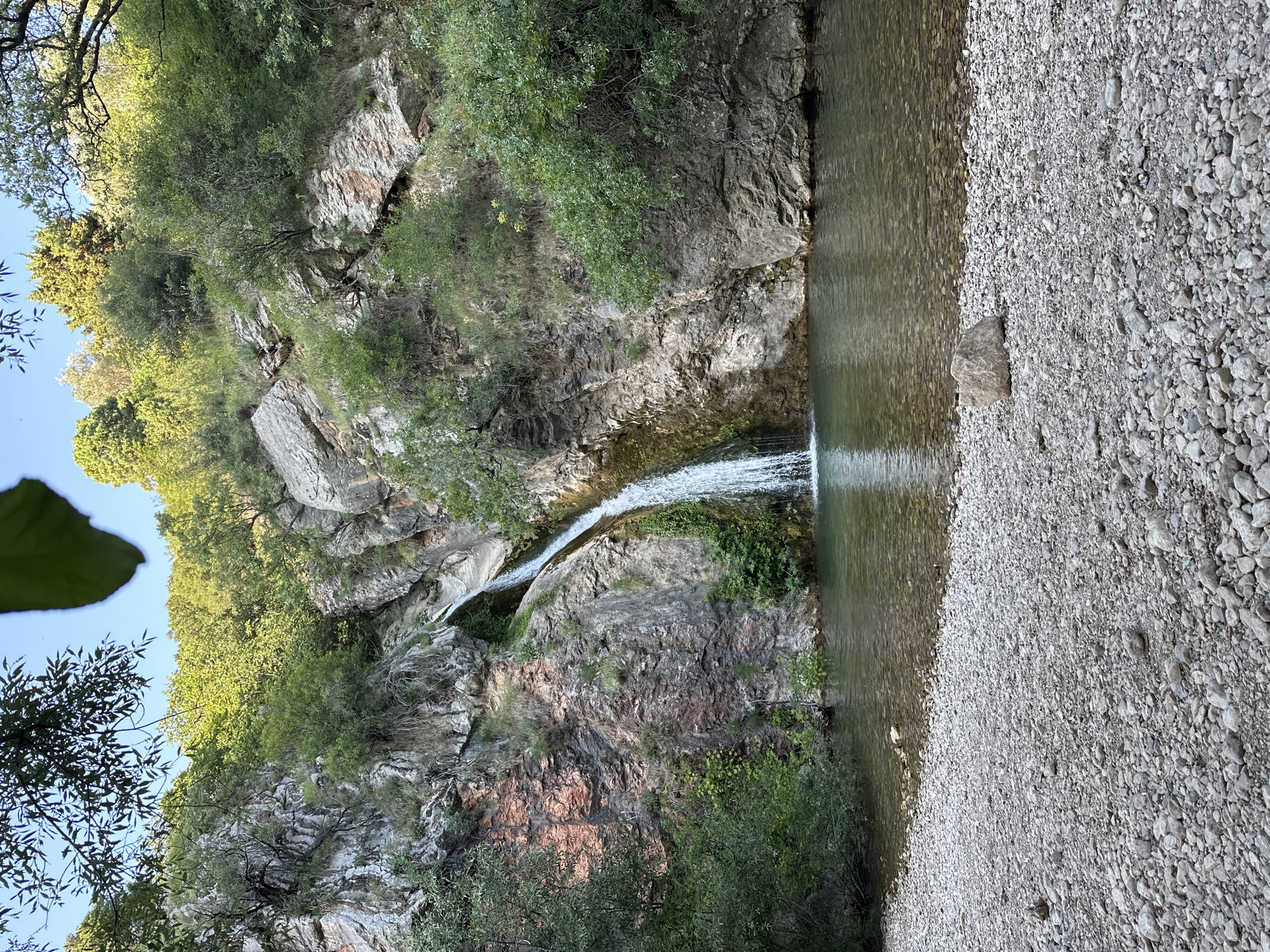 The image shows a waterfall cascading down a rocky cliff face into a clear pool of water. The surrounding environment is lush and green, with trees and vegetation growing on the slopes. The foreground is a rocky shore leading to the water's edge. The scene appears to be a natural landscape, possibly a park or nature reserve. There are no people or animals visible in the image. The location is identified as being in the city of Bečići, Montenegro.