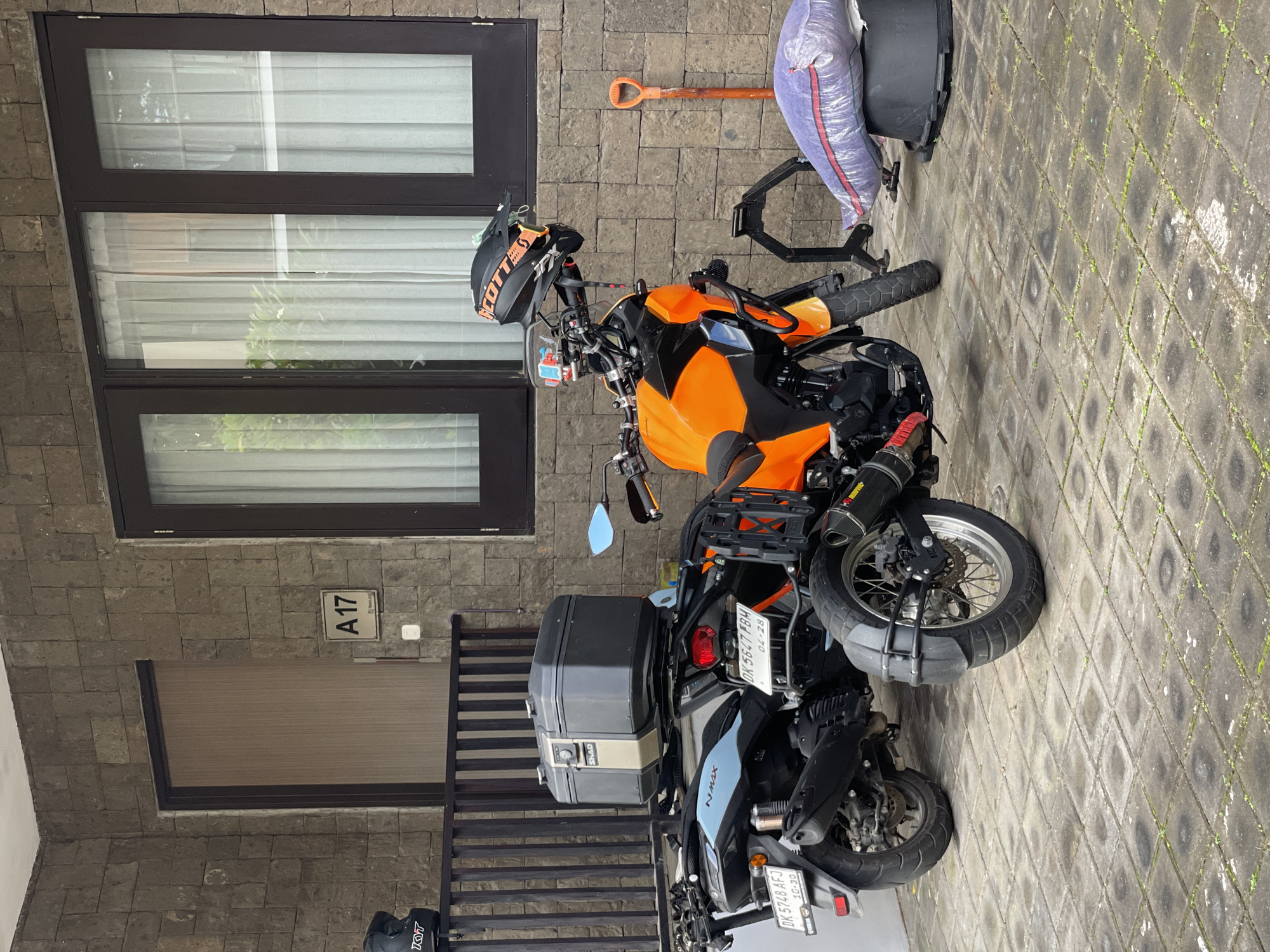 A motorcycle and a scooter are parked in front of a stone building in Kuta Selatan, Indonesia. The motorcycle is orange and black and has a helmet on the handlebars. The scooter is black and blue and has a storage box attached to the back. A shovel, a bag of supplies, and a black container are next to the motorcycle. The address "A 17" is visible on the building. The ground is paved with interlocking stone blocks.