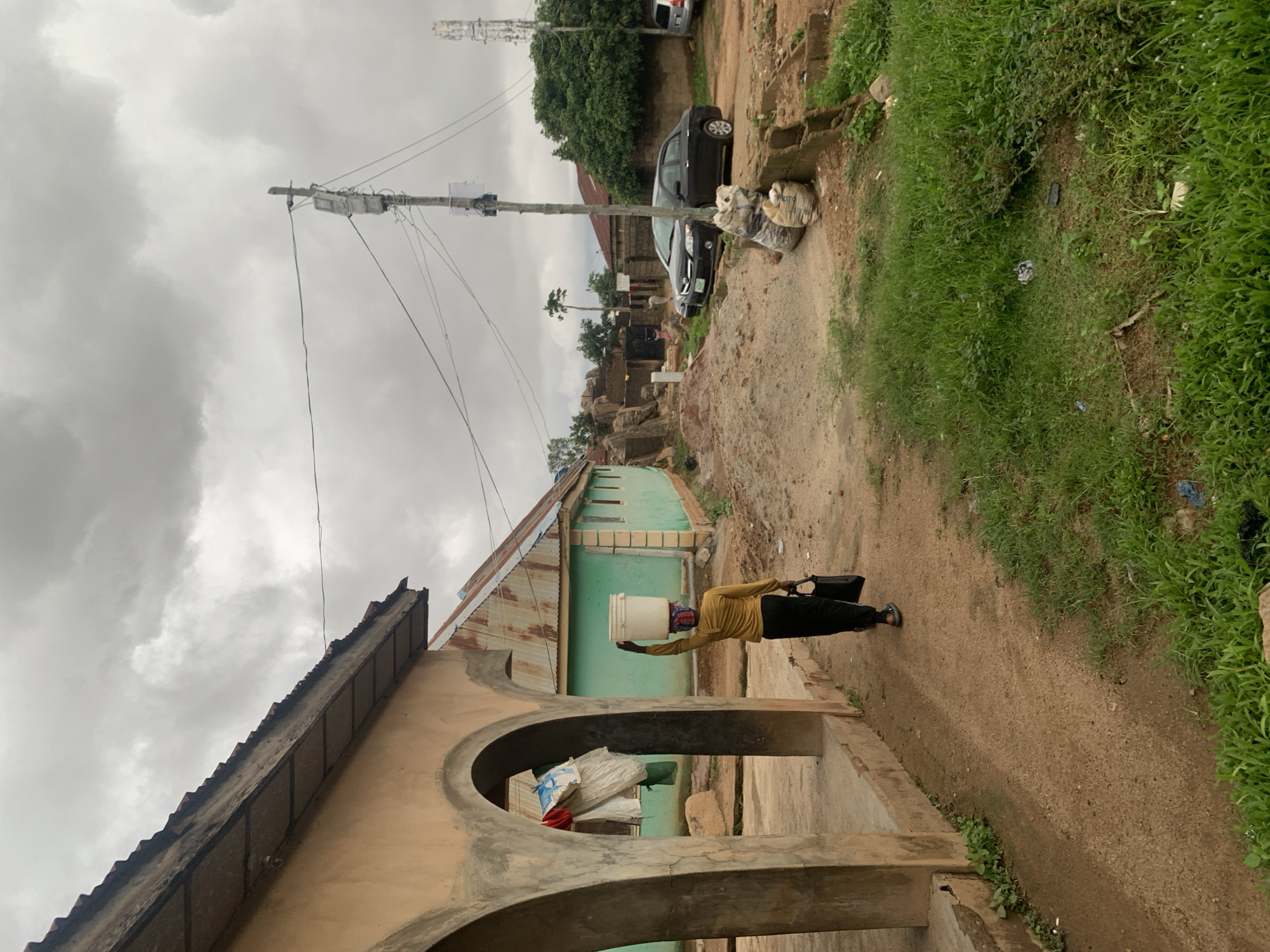 A woman walks carrying a white bucket on her head and a black bag in her hand. She is walking on a dirt path in front of a light yellow building with arched openings on the left. In the background, there is a light green building with a corrugated metal roof. There are several cars parked along the dirt road. Utility poles and wires are visible in the sky. The sky is overcast with dark clouds. The scene appears to be in a residential area in Jos, Nigeria.