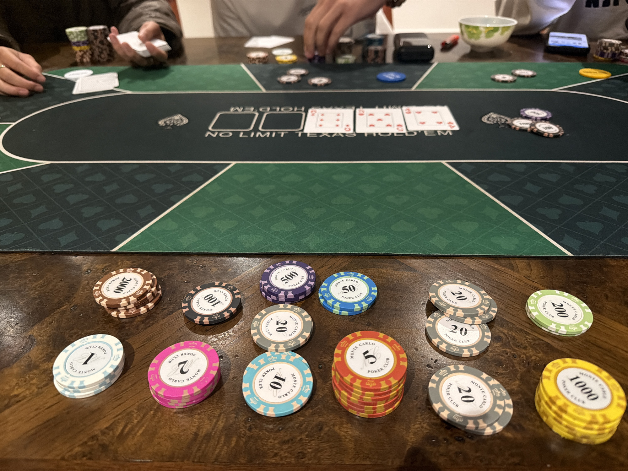 The image shows a close-up view of a poker game in progress on a wooden table. In the foreground, there are several stacks of poker chips with varying denominations and colors, indicating different values. The chips are from "Monte Carlo Poker Club" and have denominations such as 1, 2, 5, 10, 20, 50, 100, 200, 500, 1000, and 2000. In the background, a portion of the green poker table is visible with "NO LIMIT TEXAS HOLD'EM" printed on it. There are also cards and more poker chips on the table. Two individuals are partially visible, with hands engaged in the game, one holding a deck of cards. The overall environment suggests a casual or recreational setting for playing poker. The specific location within Xuân Trường - Đà Lạt, Vietnam, is not discernible from the image content.