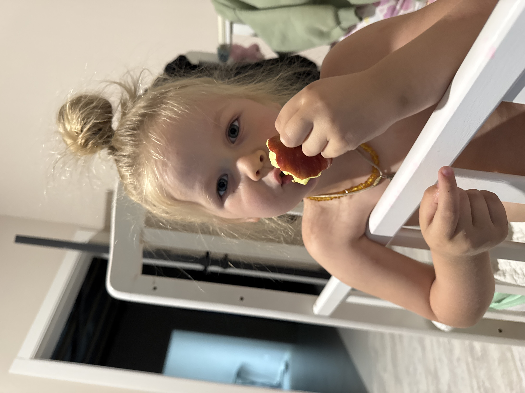 A young, blonde-haired child is eating a piece of fruit. Her hair is in a small bun on top of her head. She is wearing a beaded necklace. She is holding a white object. The location is likely indoors, and the general location is the city of Ферма, Russia.