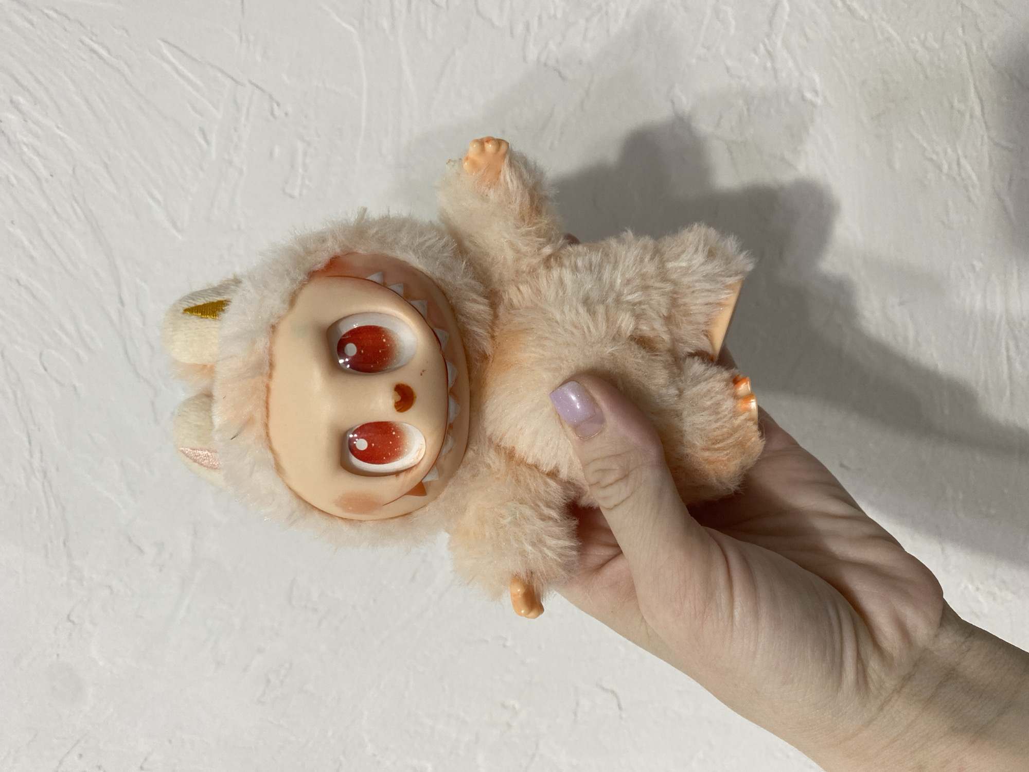 A doll-like toy is being held in a person's hand against a white textured wall. The toy is upside-down with a smooth, light-peach face that features two large red-orange eyes with white reflections, a brown dot between the eyes, and a white sawtooth design framing the face. The doll has a light-peach furry body. The hand holding the toy has pale skin and a light purple nail polish on the thumb. The background is a white wall with a rough, textured surface. The location is not provided.