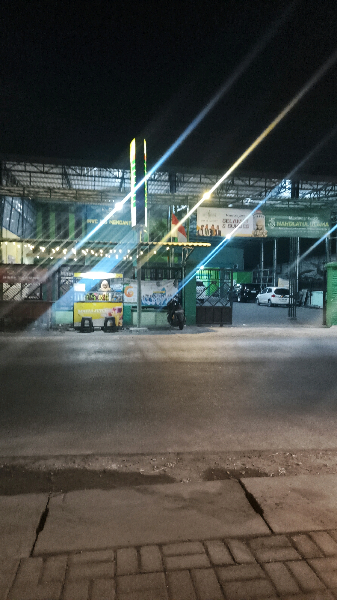The image captures a nighttime scene outside a building with a green facade in Pelemwatu, Indonesia. The building has a covered area with numerous string lights illuminating the front. In the foreground, a paved road is visible with a sidewalk made of interlocking bricks.
To the left, a mobile food cart is set up. It is yellow and decorated with images of fruits and the text "MARTA JUICE." Two stools are placed in front of the cart. A person wearing a headscarf is visible behind the counter of the food cart.
A motorcycle is parked to the right of the food cart. Beyond the food cart and motorcycle, a gate is visible. Through the gate, cars can be seen parked.
Banners are displayed on the building. One banner, partially visible on the left, has text that appears to include "MWC MENGANT." Another banner to the right contains text including "Selamat" and "Muktamar Ke-35 NAHDLATUL ULAMA." The building's structure suggests it might be a community hall or educational facility, given the banners referencing "Muktamar" (congress). The lighting creates strong light streaks due to the camera's exposure settings.
