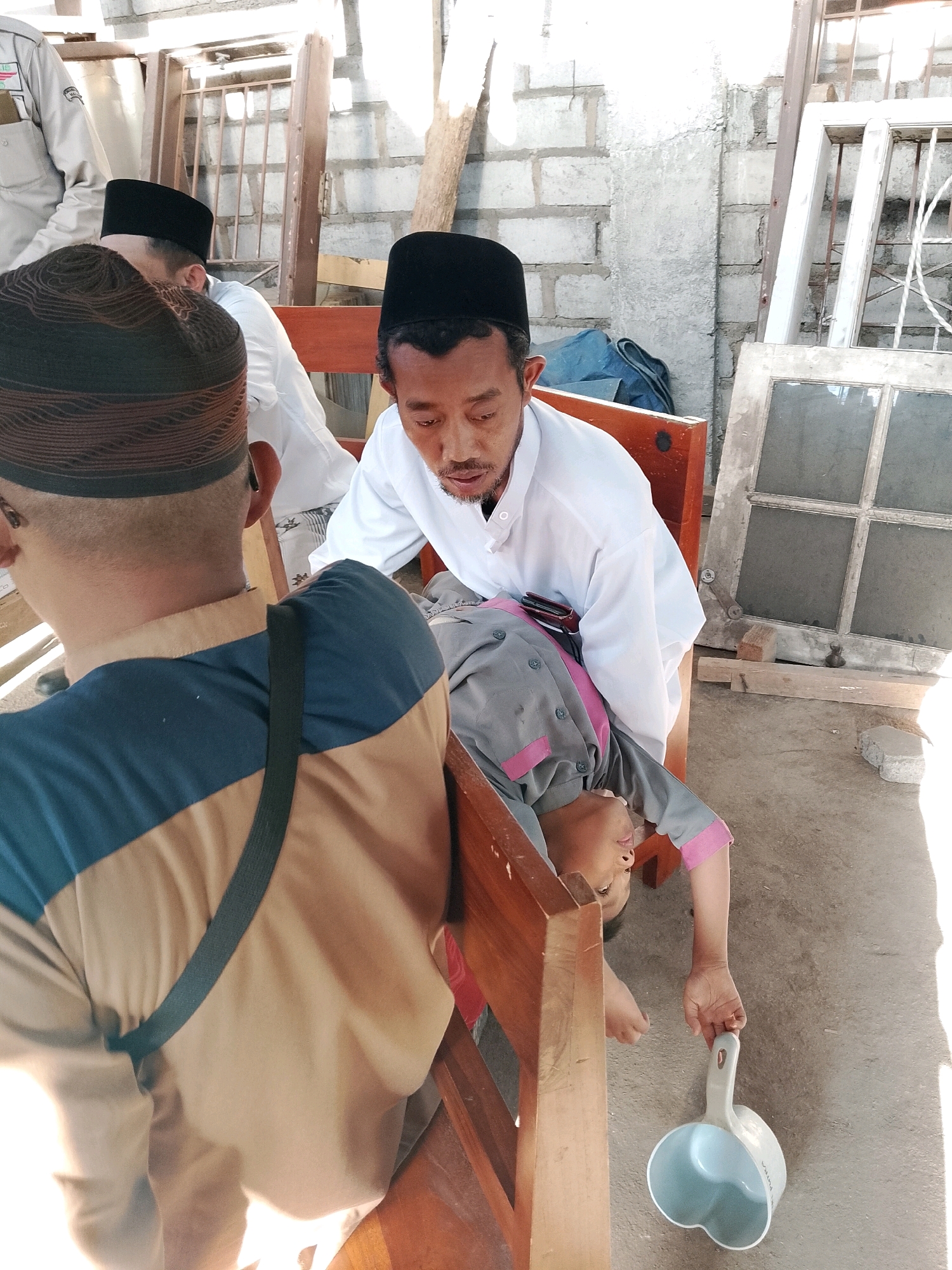 The image depicts a scene in Banjarejo, Indonesia, with several individuals present. In the foreground, the back of a person wearing a brown and blue garment with a black strap is visible. To their right and slightly further back, a man in a white garment and black cap is leaning over another person. This second person, dressed in a grey and pink uniform, is lying down or reclined on a wooden bench, with their head tilted back. They are holding a light-colored ladle in their right hand. In the background, other individuals can be seen, also wearing caps. The environment appears to be an outdoor or semi-outdoor area, possibly a courtyard or a workspace, with brick walls, wooden structures, and discarded window frames visible. The ground is a concrete surface. The lighting suggests it is daytime.