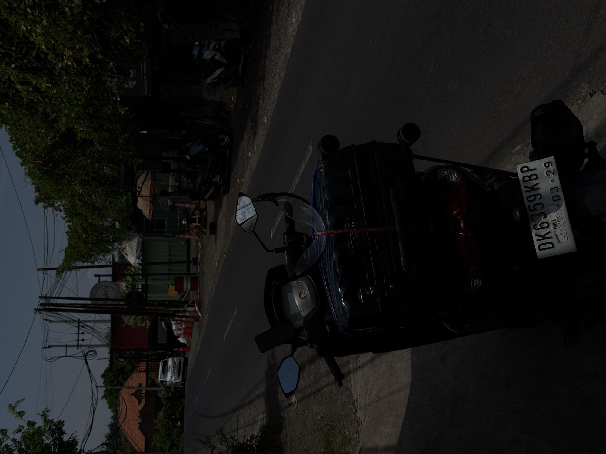 A dark-colored Yamaha NMAX scooter is parked on the left shoulder of a paved road in Desa Belalang, Indonesia. A black hard-shell suitcase is secured to the rear seat with blue and red straps. A clear helmet visor rests on the handlebars, and blue-tinted rearview mirrors are visible. The rear license plate reads "DK 6359 KBP" with "03 29" below it, and "THEA Scooter Rental" is printed on a sticker underneath the plate. The road ahead shows faint white lane markings and tire scuffs on the asphalt.
In the midground, to the left, a traditional Indonesian building with an orange tiled roof is visible behind dense foliage. A white passenger van is parked further down the road, and several orange traffic cones are positioned near a red drum, possibly a Pertamina fuel barrel or barrier. Numerous overhead electrical wires are strung between utility poles throughout the scene.
To the right, a dense canopy of green trees lines the roadside. Another dark-colored scooter is parked beneath the trees. A commercial building, partially obscured by trees, features a circular white sign with "Rigby's" displayed and another dark rectangular sign beneath it. An orange umbrella is visible further down the road, near another parked scooter. The sky above is bright blue with scattered clouds, indicating daytime conditions.