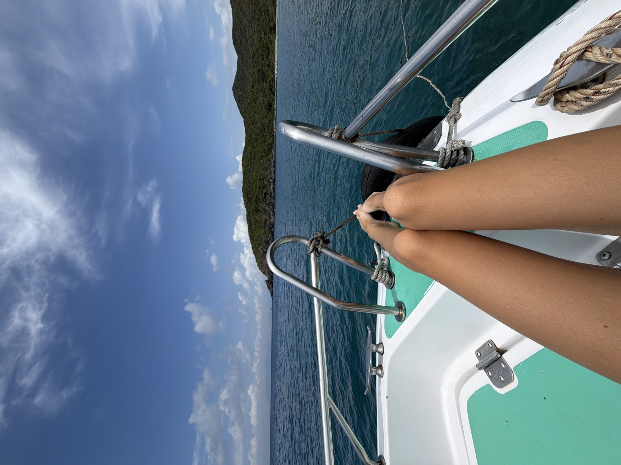 Aboard a boat sailing on the water in Nha Trang Ward, Vietnam, a person's legs are visible, tanned, resting on the deck of the boat. The deck is white with patches of green. Stainless steel railing is visible. The rope is tied to a cleat. A coastline is visible in the distance, covered in greenery. The sky is a bright blue with wispy clouds. The water stretches out to the horizon.
