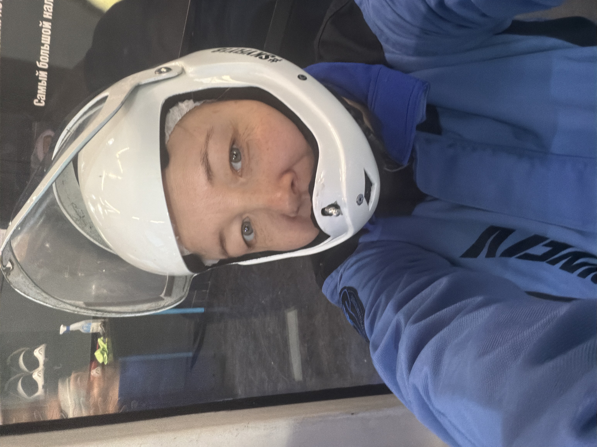 A woman wearing a white helmet with a clear visor and a blue jumpsuit is posing for a selfie. The helmet appears to have the word "#SKYDIVING" on its side. She is inside what appears to be a wind tunnel facility. Reflections visible in the background and visor indicate the presence of signage in Russian. One sign includes the phrase "Самый большой нал". Based on the jumpsuit and helmet, the subject is likely engaged in or preparing for indoor skydiving. The location is Izhevsk, Russia.