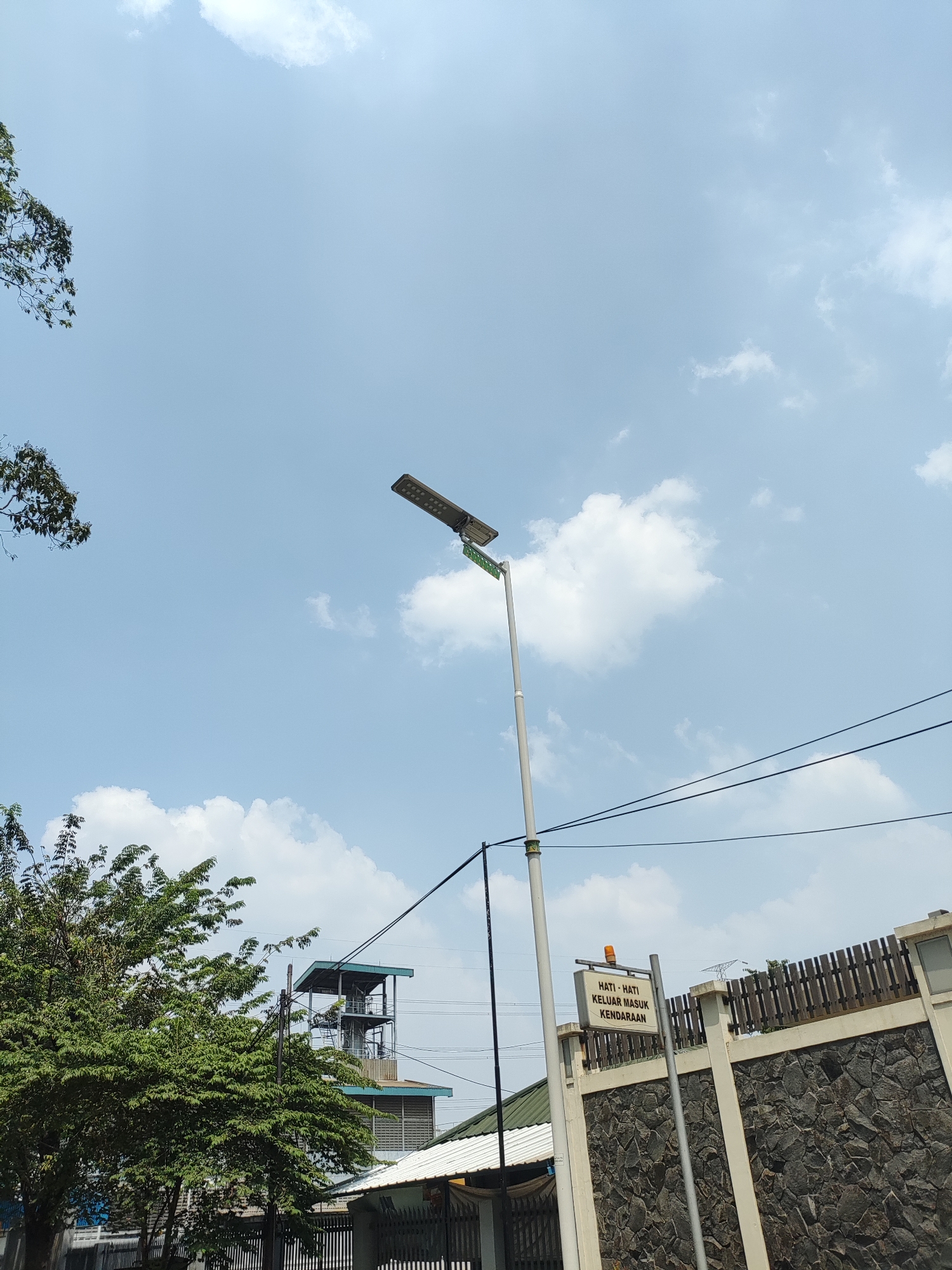 This is a photograph taken on a clear day in East Jakarta, Indonesia, showing a street lamp and a warning sign. The street lamp, which appears to be a solar-powered LED lamp, is a prominent feature in the upper center of the image, with its solar panel visible at the top. Below the lamp, a white sign with orange text reads "HATI - HATI KELUAR MASUK KENDARAAN," which translates to "CAUTION - VEHICLE ENTRANCE AND EXIT." This sign is mounted on a pole and is positioned to the right of the street lamp.
The background reveals a mix of urban and natural elements. A dense green tree with many leaves is located in the lower left quadrant of the image, its branches extending upwards towards the sky. Behind the tree and to the right, a multi-story building with a metallic structure and what appears to be a control tower or observation deck can be seen. Further to the right, a wall constructed with dark, irregular stones forms the foreground. Above the stone wall, a wooden fence with vertical slats runs along the top. Power lines are strung across the sky, crossing diagonally from the left to the right side of the frame. The sky itself is a bright blue, punctuated by scattered white clouds. The overall scene suggests a street or an entrance to a facility in an urban environment.