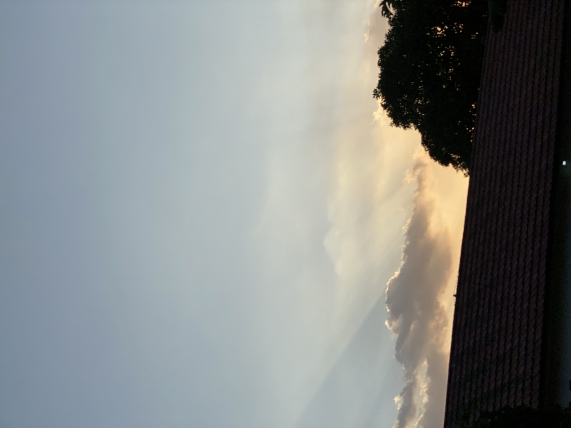 An overcast sky with crepuscular rays is visible over a tiled roof and silhouetted trees. The location is Banjarbaru, Indonesia. The image appears to have been taken during sunset, with the sky displaying a gradient of light blue to pale yellow. The clouds are dimly lit, and the rays of sunlight are piercing through them. The roof in the foreground is dark red and has a tiled pattern. A large, dark tree is silhouetted against the sky on the right side of the image. The environment is urban or suburban, indicated by the presence of a roof and trees. There are no people or animals visible in the image. The overall atmosphere is calm and serene.
