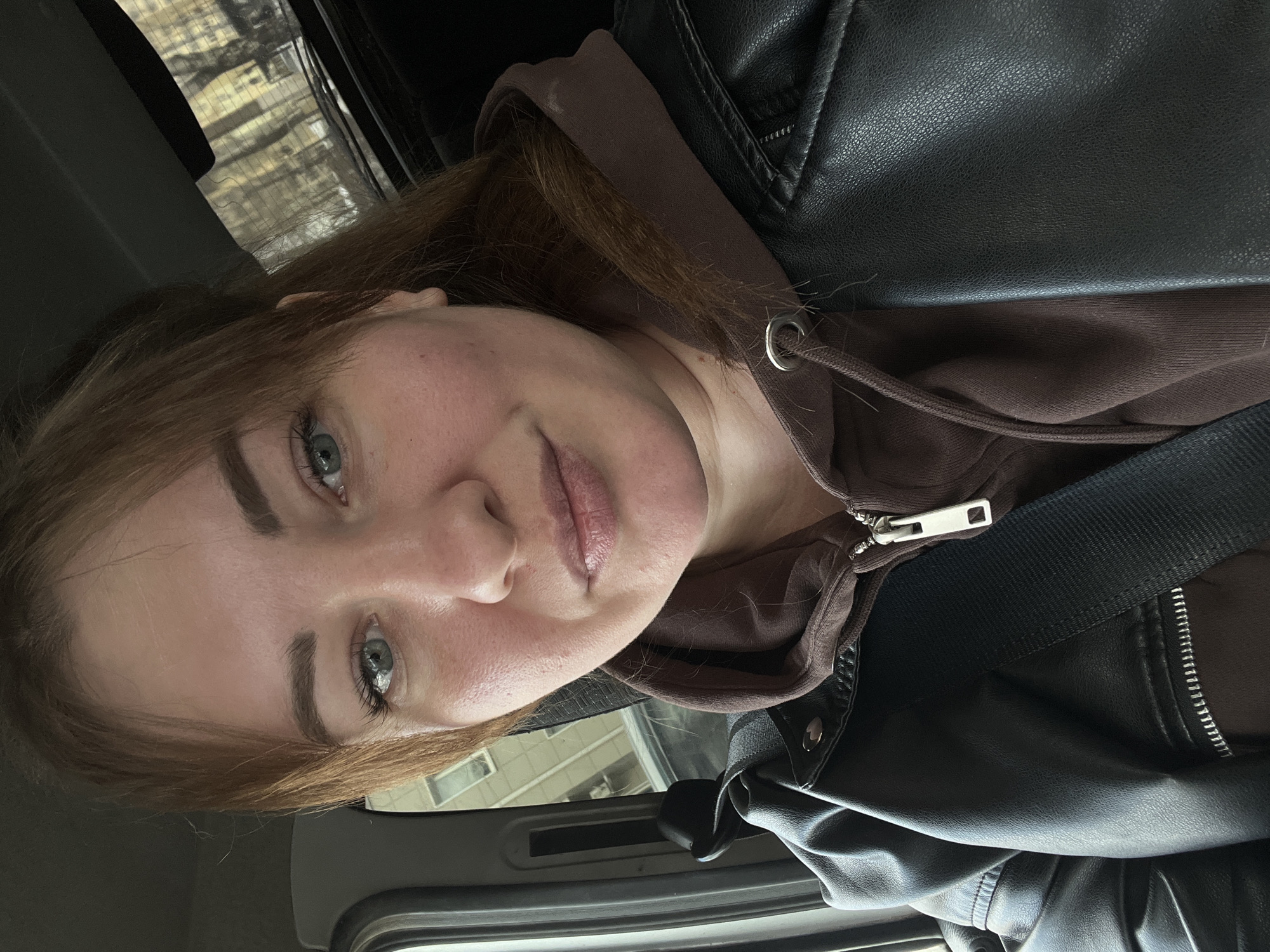 A female individual is depicted in a close-up portrait, seated inside a vehicle. Her light brown hair, styled with bangs, frames her face, revealing light blue-green eyes. She wears a brown hooded sweatshirt beneath a black faux leather jacket with a visible zipper and collar snaps. A black seatbelt is fastened across her chest. The vehicle's grey interior trim is visible to her left. Through the left window, a light-colored multi-story building with rectangular windows is partially visible. Through the rear window, a blurred view of leafless trees against a lighter background suggests an urban environment. No specific landmarks are present to determine a precise location within the city of Chaikovsky, Russia.