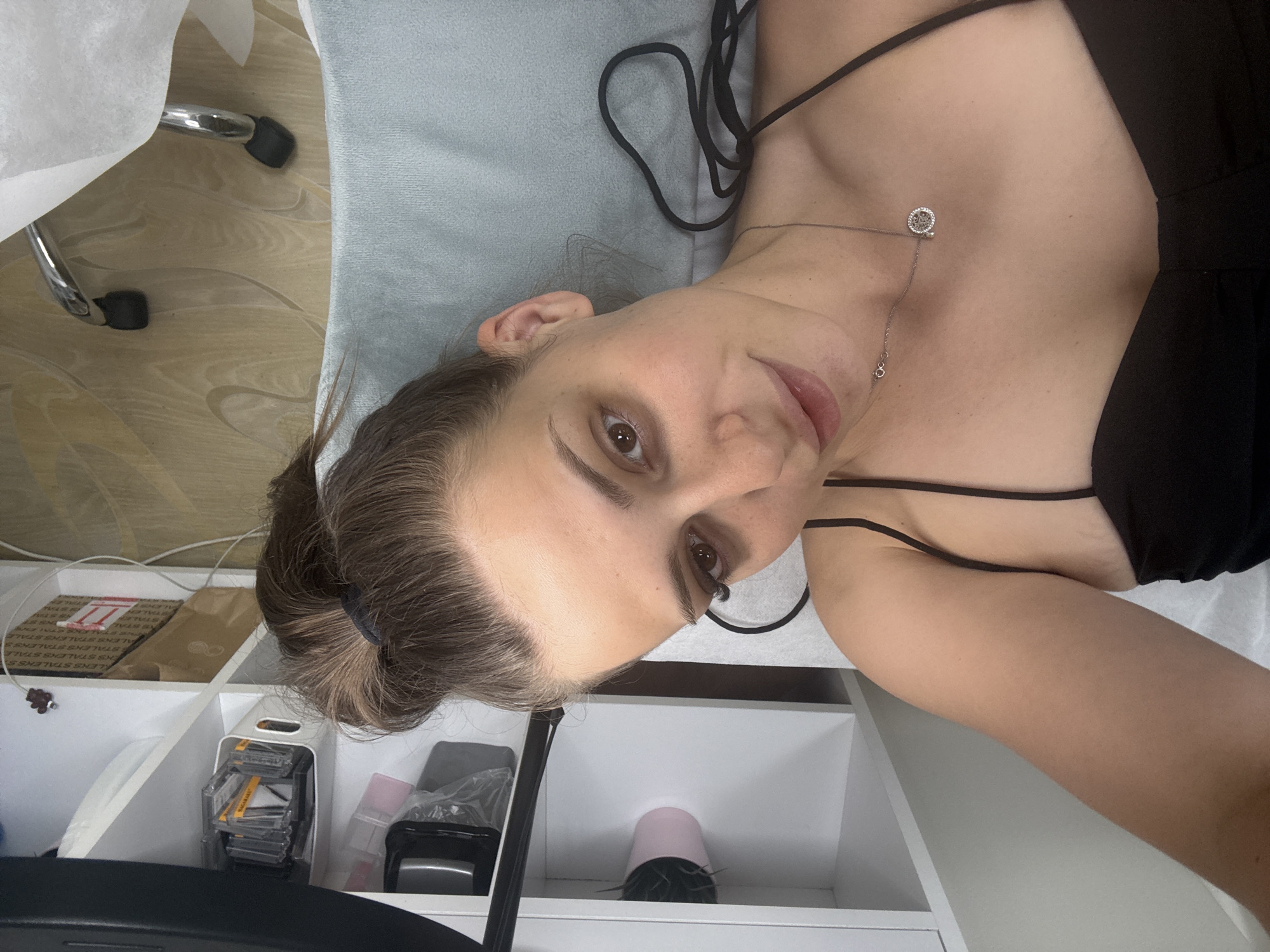 A woman with her hair in a bun lies on a light blue surface, looking at the camera. She is wearing a black camisole and a silver necklace with a circular pendant. The background shows white shelving units filled with various items, including what appear to be beauty products and boxes. A rolling chair with chrome legs and a white cover is visible in the upper right corner. The scene appears to be an indoor setting, possibly a salon or treatment room. The location is not specified in Plovdiv, Bulgaria.