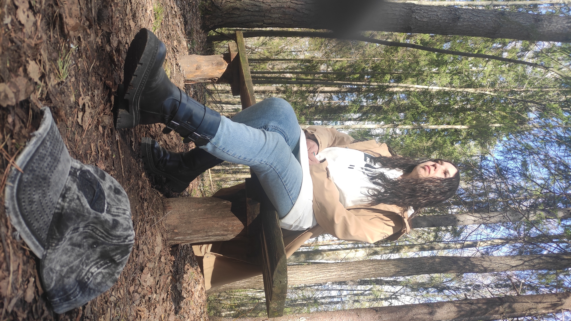 The image shows a woman sitting on a wooden structure resembling a bench in a wooded area. The woman is wearing a tan coat, a white t-shirt, light blue jeans, and black boots. A grey denim cap is visible on the ground near her feet. The environment consists of trees, foliage, and fallen leaves. The scene appears to be outdoors in a forest or park setting, although Noviy, Russia is not discernible from this image.