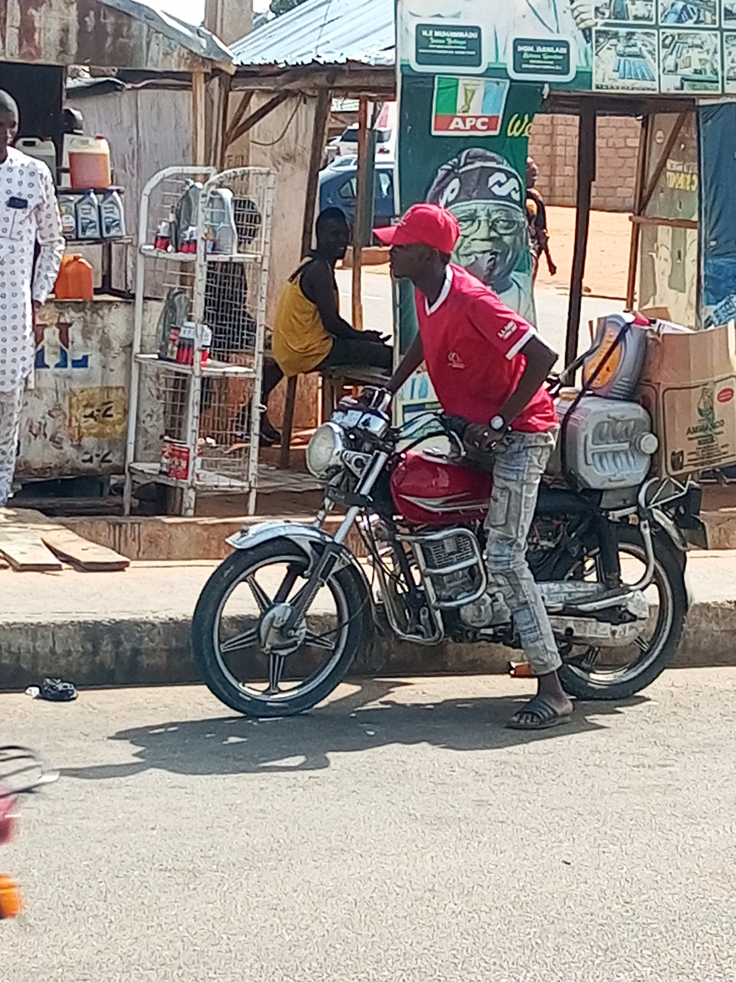 The image captures a bustling, sunlit street scene in Garko, Nigeria, featuring several individuals and roadside stalls. The bright daylight casts distinct shadows, indicating clear weather.
In the foreground, a young man wearing a red t-shirt, light-colored patterned jeans, and a red baseball cap sits astride a red motorcycle. He is wearing black slides on his feet. The motorcycle is heavily loaded with goods, including two large, light-grey plastic containers and a cardboard box labeled "AMIRADO" secured to the rear, along with a prominent yellow plastic jerry can. The man appears to be paused, perhaps at a stop or preparing to depart, suggesting he might be a delivery rider or a vendor.
To the left, partially visible, is a man in a white and black patterned long-sleeved garment, standing near a makeshift stall. This stall, constructed from metal racks and other materials, displays an assortment of goods, including multiple plastic bottles of motor oil and at least two orange jerry cans, suggesting it functions as a lubricant or fuel station. Further back on the right, another man, dressed in a yellow tank top, is seated on a stool behind a similar, less visible stall structure.
In the background, a large green political banner is prominently displayed. It features a portrait of a man, likely a political figure, and the clear "APC" (All Progressives Congress) logo, a major Nigerian political party. Visible text on the banner includes "N.F. MUHAMMADU" and "HON. BAYANLI" (partially obscured). The surrounding environment includes simple structures with corrugated metal roofs and various wall materials, hinting at local businesses or residences along a paved road with a raised curb.