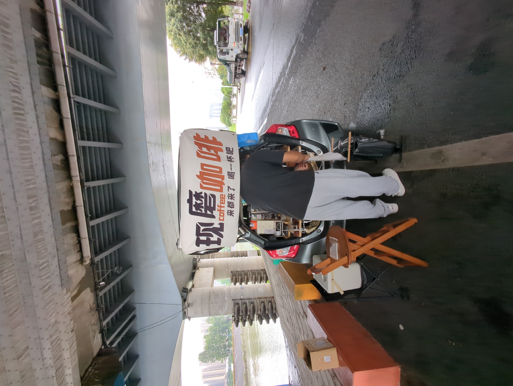 A street-level view under an overpass shows a person operating a mobile coffee station set up in the back of a gray vehicle. The person, wearing a dark shirt and light gray pants, is facing the open hatchback. A large sign above the vehicle reads "现磨咖啡" in Chinese characters, with "Coffee" in English underneath. The area around the vehicle is wet, suggesting recent rain. A folding wooden chair and a white cooler are near the vehicle. A river is visible behind the overpass support structures, and a city skyline is in the background. Other vehicles are parked further up the road. The scene appears to be in the Shunde District of China, based on the Chinese characters on the sign and the general appearance of the environment.