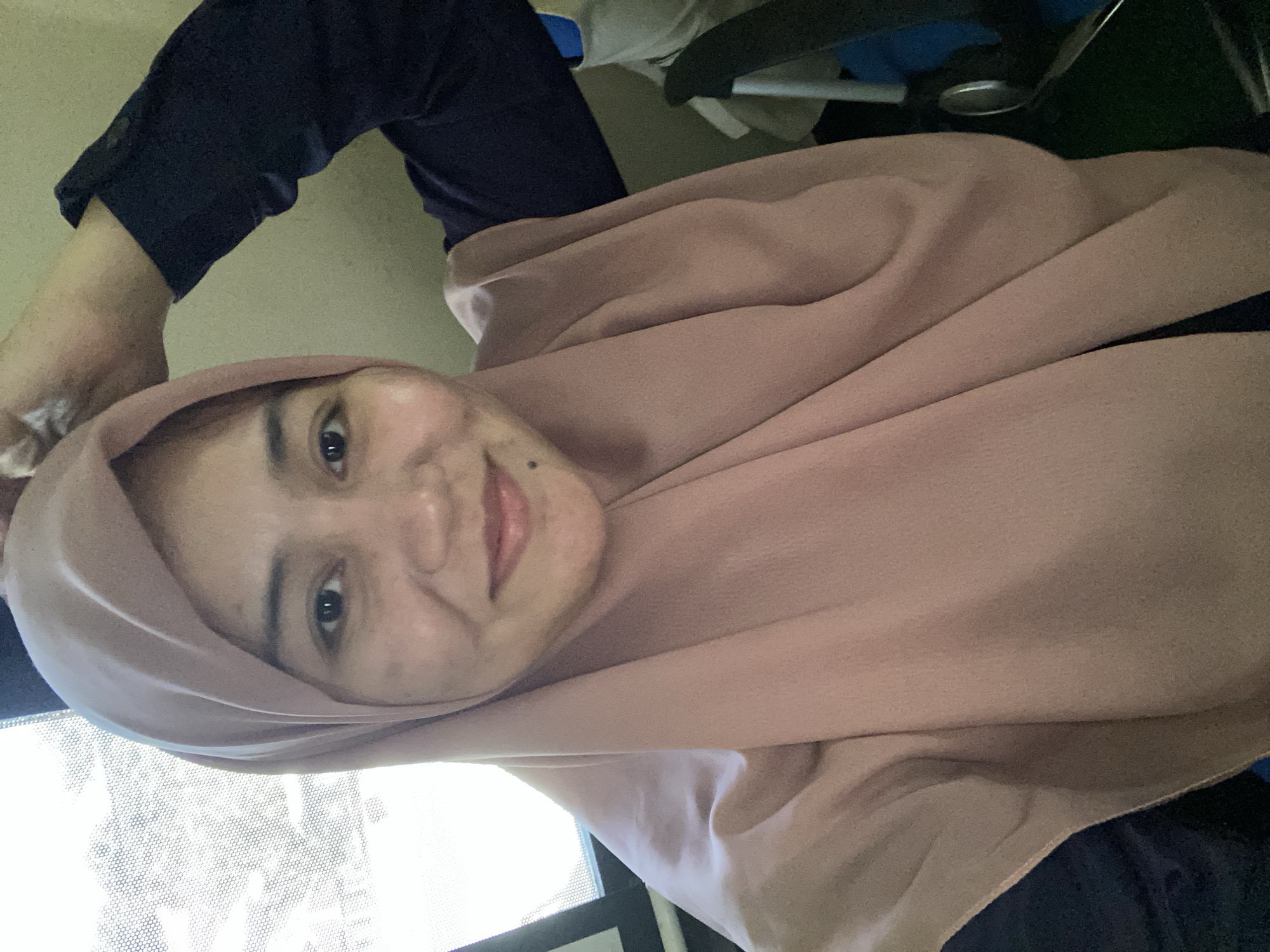 A young Indonesian woman wearing a pale pink hijab and a dark blue long-sleeved shirt smiles at the camera. Her right hand is raised and touches the top of her head. The background consists of a window with a patterned screen to the left, and a wall and office chair to the right. The scene appears to be indoors, possibly in an office or a room in Banjarbaru, Indonesia, given the context. The lighting suggests it is daytime, with natural light coming from the window.