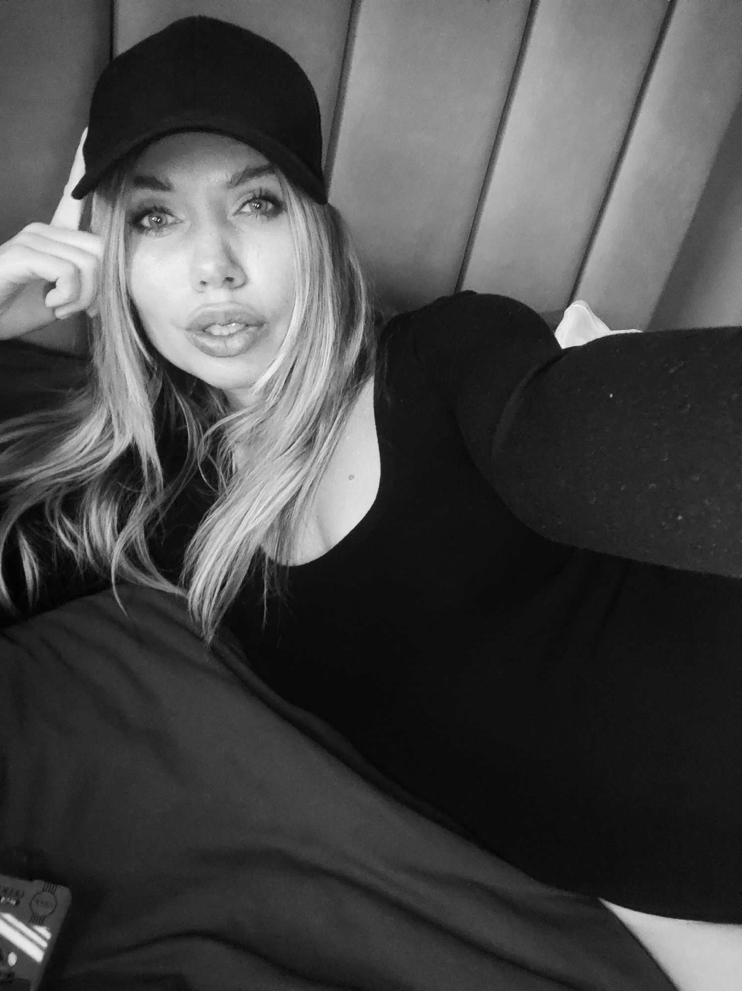 This black and white portrait features a woman posing for a close-up selfie, likely taken indoors in Votkinsk, Russia. The woman has long, flowing, light-colored hair and is wearing a dark (appearing black in monochrome) baseball cap with the brim pointed forward, along with a dark long-sleeved top or dress. Her right hand is raised, with her fingers gently resting against her temple and the side of her cap.
She looks directly into the camera with prominent eyes and a neutral to slightly pensive expression, her lips slightly parted. The background consists of a padded or upholstered headboard or wall paneling, with vertical sections visible in varying shades of gray. Part of a white object, possibly a pillow or blanket, is visible behind her right arm. In the bottom left corner, an indistinct object with some blurry white markings and symbols, possibly a mobile phone, is partially visible, but no clear text is legible. The lighting appears soft, creating a direct and engaging composition.