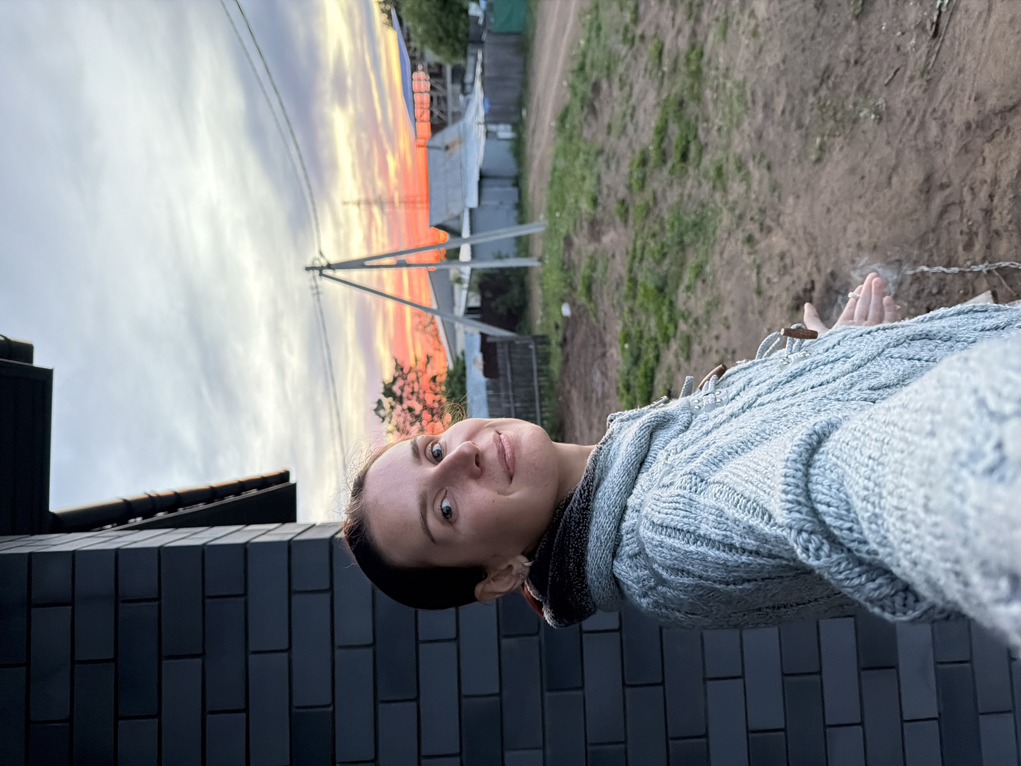 A woman is pictured in a vertical selfie against a backdrop of a sunset and a rural landscape. The woman has fair skin, short dark hair, and is wearing a light blue knitted sweater. She is smiling slightly at the camera. Behind her, there is a dark-colored brick wall, likely part of a building. Beyond the wall is a view of utility poles and wires silhouetted against a colorful sunset sky with hues of orange, pink, and grey. In the distance, there are modest houses and vegetation suggestive of a rural or suburban setting. The ground appears to be a mix of dirt and grass. In the bottom right corner, the woman’s hand holding a smoldering cigarette is partially visible. Given the mention of the city of Chaikovsky, Russia, it can be inferred that the location is in or near this city, although specific landmarks are not visible in the frame to confirm the exact location.