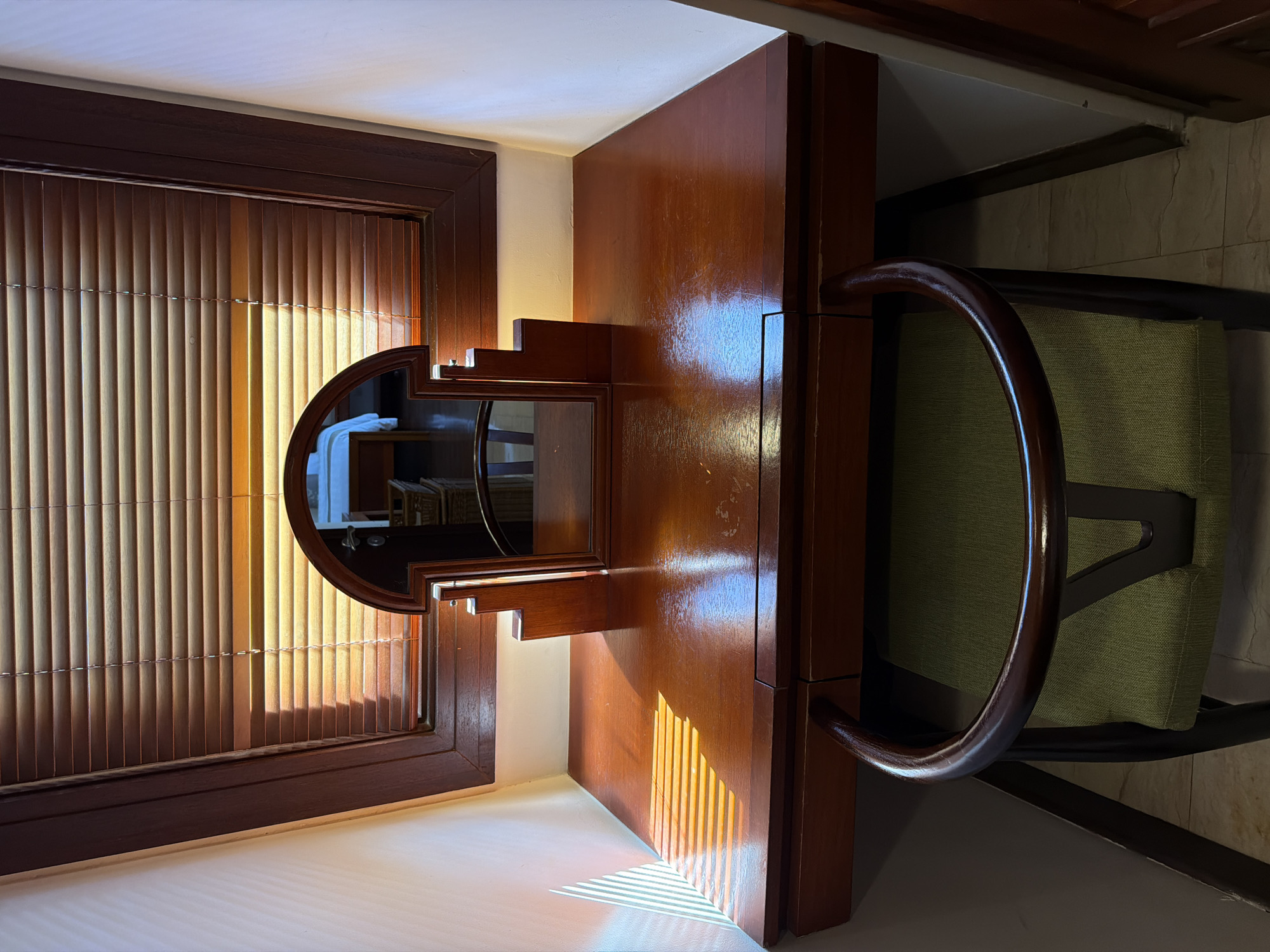 This image, captured in Kuta, Indonesia, presents a sunlit interior space, likely a hotel room or bedroom, characterized by warm wooden furnishings and natural light. The primary subjects are a dark wooden dressing table or desk and a corresponding chair, set against a window.
On the left, a large window is fitted with vertical blinds, possibly made of wood or bamboo, allowing warm sunlight to penetrate the room. This light creates striking diagonal stripes of illumination and shadow across the polished surface of the wooden table and the lighter-colored surface beneath the window, indicating a late morning or early afternoon time of day. The window itself is framed in dark wood.
The dressing table features a round mirror on its left, also encased in dark wood, with a distinct tiered, stepped architectural detail on one side. The mirror's reflection offers a peek into the opposite side of the room, revealing what appears to be white fabric or a towel, and potentially shelving or baskets.
To the right of the table stands a chair, constructed from the same dark wood, with a gracefully curved armrest that integrates into its back frame. The seat and backrest of the chair are upholstered in a light green, textured fabric. The floor visible near the chair is tiled in a neutral, light hue.
The scene is calm and static, depicting no specific activity. The dominant aesthetic is one of rich wood tones bathed in natural sunlight. No text is visible in the media file.