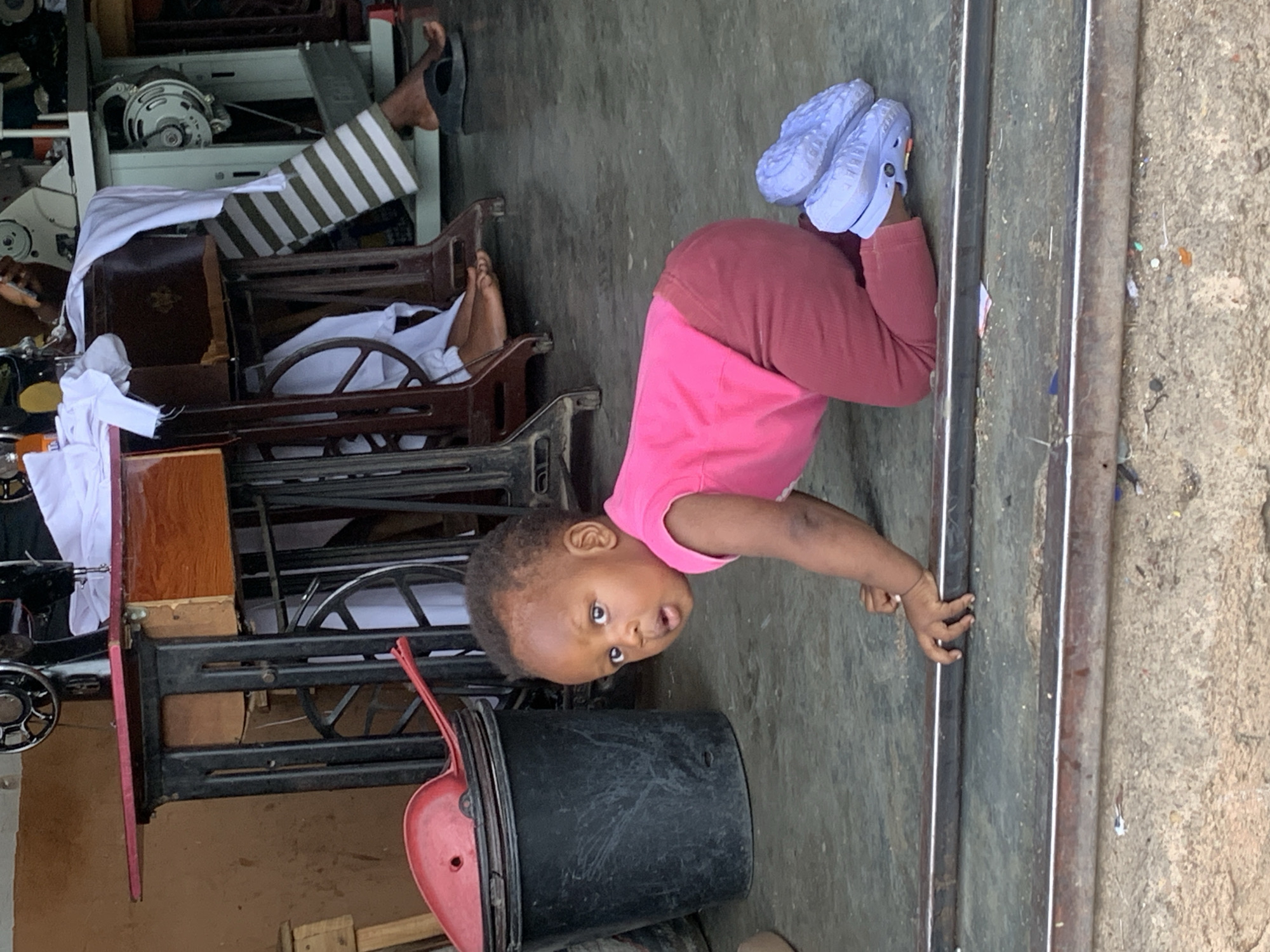 A toddler wearing pink clothing and blue shoes is crawling on a concrete floor indoors. They are positioned to the right of a metal rail or track. To the left, there are several antique sewing machines stacked together. A black bucket with a red scoop sits in front of the sewing machines. In the background, there is a person reclining in one of the sewing machines. This image was taken in Jos, Nigeria.