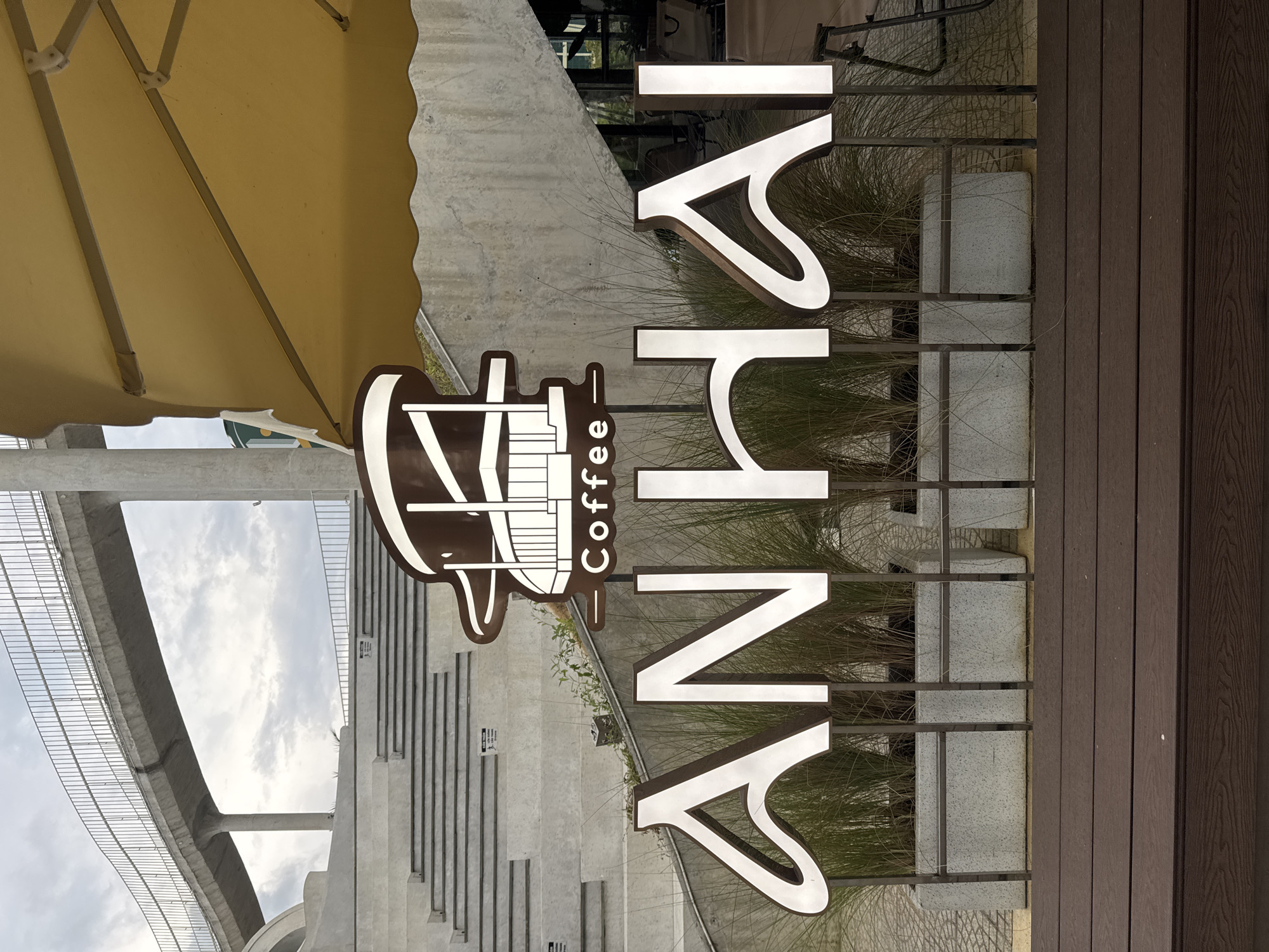 The image shows a sign for "AnHai Coffee" in Đà Nẵng, Vietnam. The sign features a stylized coffee cup graphic next to the vertical text. The background includes stairs leading up to a building and an elevated bridge structure. There is a brown, wooden deck or walkway to the right of the sign and vegetation is visible behind it. A yellow shade is partially visible in the top left corner. The sky is overcast.