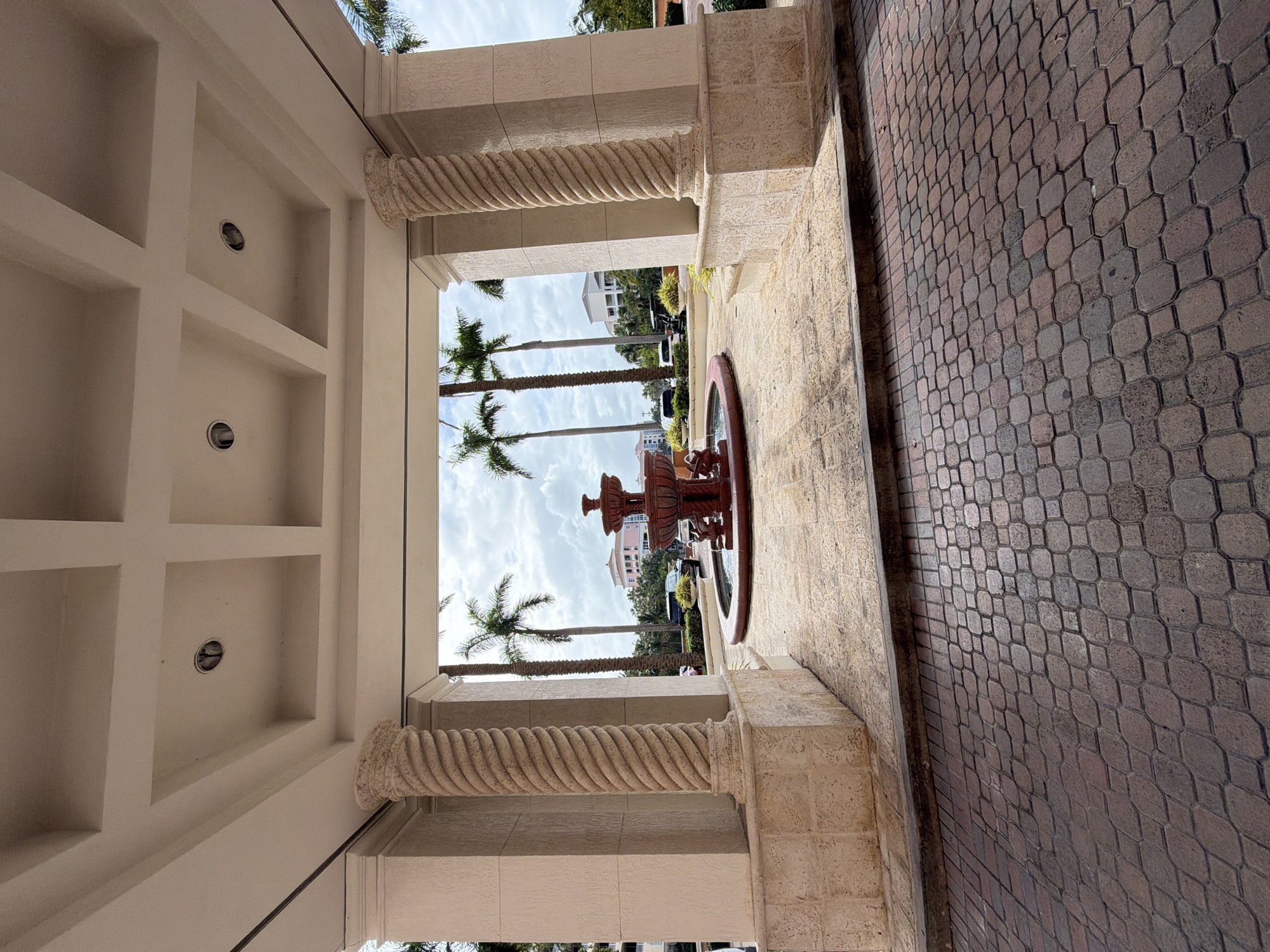 This image shows a view through an arched entranceway in Key Biscayne, Florida, USA. The entranceway is framed by two decorative columns with a spiral design, supporting a coffered ceiling with recessed lighting. Beyond the entrance is a courtyard with a tiered fountain in the center, surrounded by palm trees and other greenery. In the background, buildings are visible. To the right of the entrance is a paved walkway with a patterned brick design. The weather appears partly cloudy, with the sky visible between the palm trees.