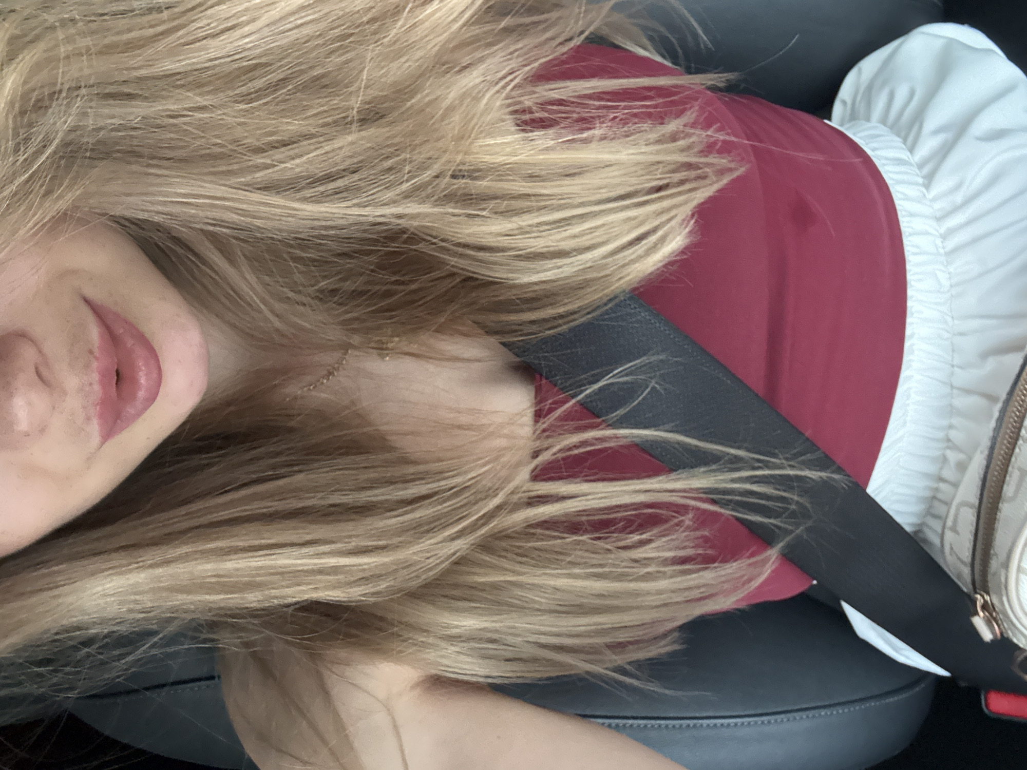 An up-close, medium shot captures the upper body of a young woman with blonde hair in a car. She is wearing a maroon tank top and white pants. A black seatbelt crosses her chest, and a portion of a light-colored handbag is visible at the bottom of the frame. Her mouth is slightly open, revealing pink lips, and her blonde hair frames her face. The background consists of dark car seats and a dark car interior. The image appears to be taken from a slightly low angle, looking up at the woman.