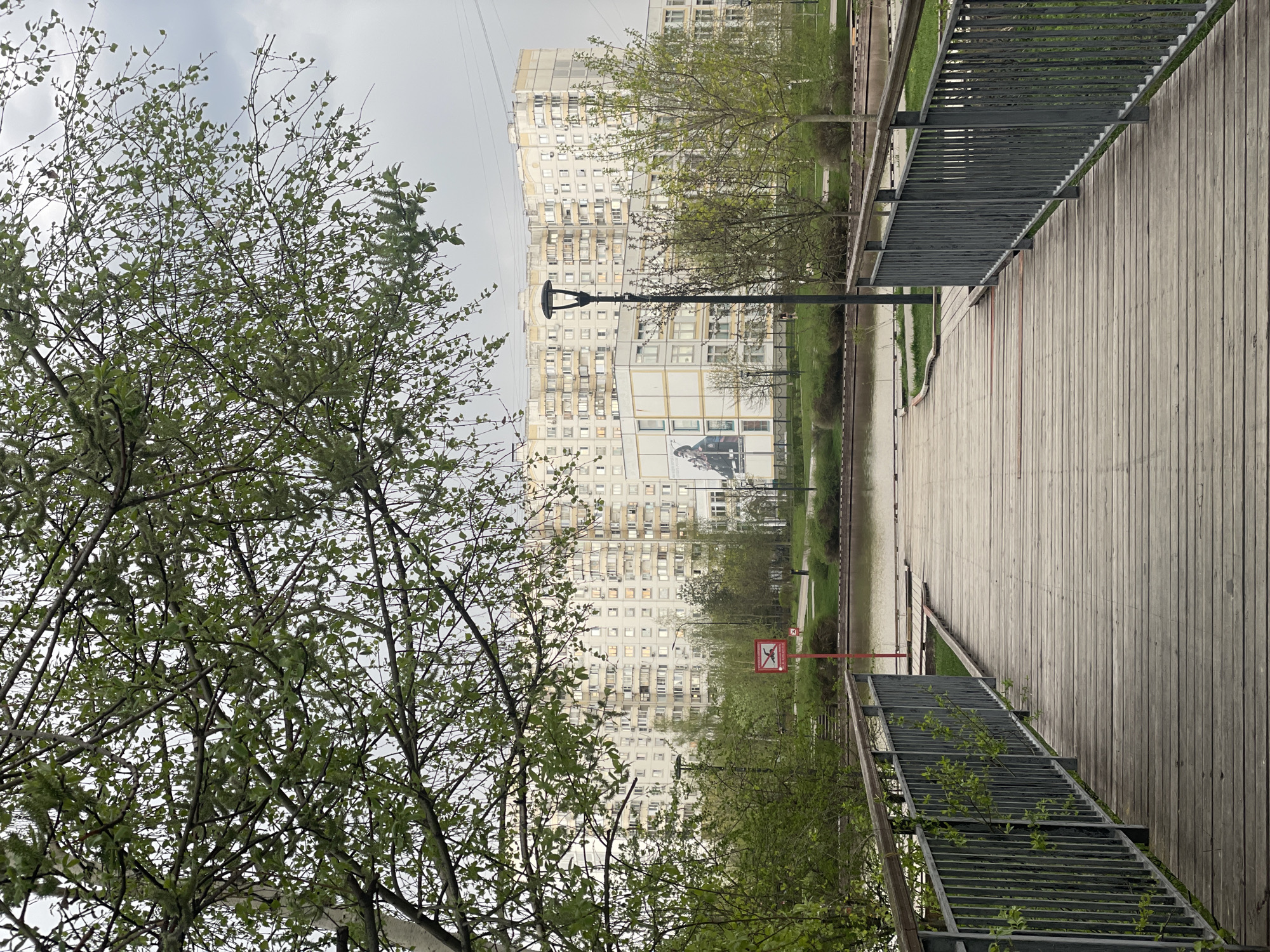The image depicts a cityscape in Moscow, Russia, dominated by a large residential building with numerous windows. A wooden boardwalk extends into the scene, flanked by metal railings and greenery. The presence of young trees with fresh leaves suggests it is spring. A red sign is visible near the boardwalk, and a lamppost stands in the middle ground. The sky is overcast, casting a diffused light over the scene. No people are visible.