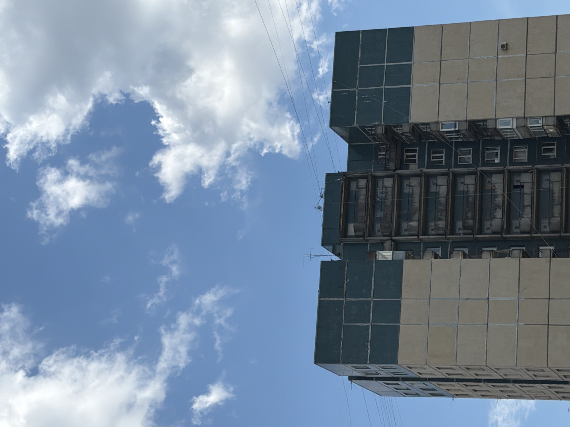 The image displays a multi-story residential panel building against a clear blue sky filled with scattered white cumulus clouds. The building's facade is segmented into large rectangular panels, featuring a dark greenish-blue color on the upper tiers and a faded beige-tan on the lower sections, with visible vertical and horizontal panel joints. The structure has a complex shape with projecting and recessed sections; a central recessed area is characterized by multiple stacked balconies, some open and some enclosed with varying materials. Rectangular windows are distributed across the visible parts of the building. On the roofline, several television antennas are present. Thin utility cables traverse the upper portion of the frame horizontally, crossing the sky. A small, light-colored rectangular unit, possibly an air conditioner, is mounted on the beige facade of the rightmost building section. No individuals, vehicles, or explicit ground-level features are visible. This architectural style is characteristic of residential construction in the city of Saint Petersburg, Russia.