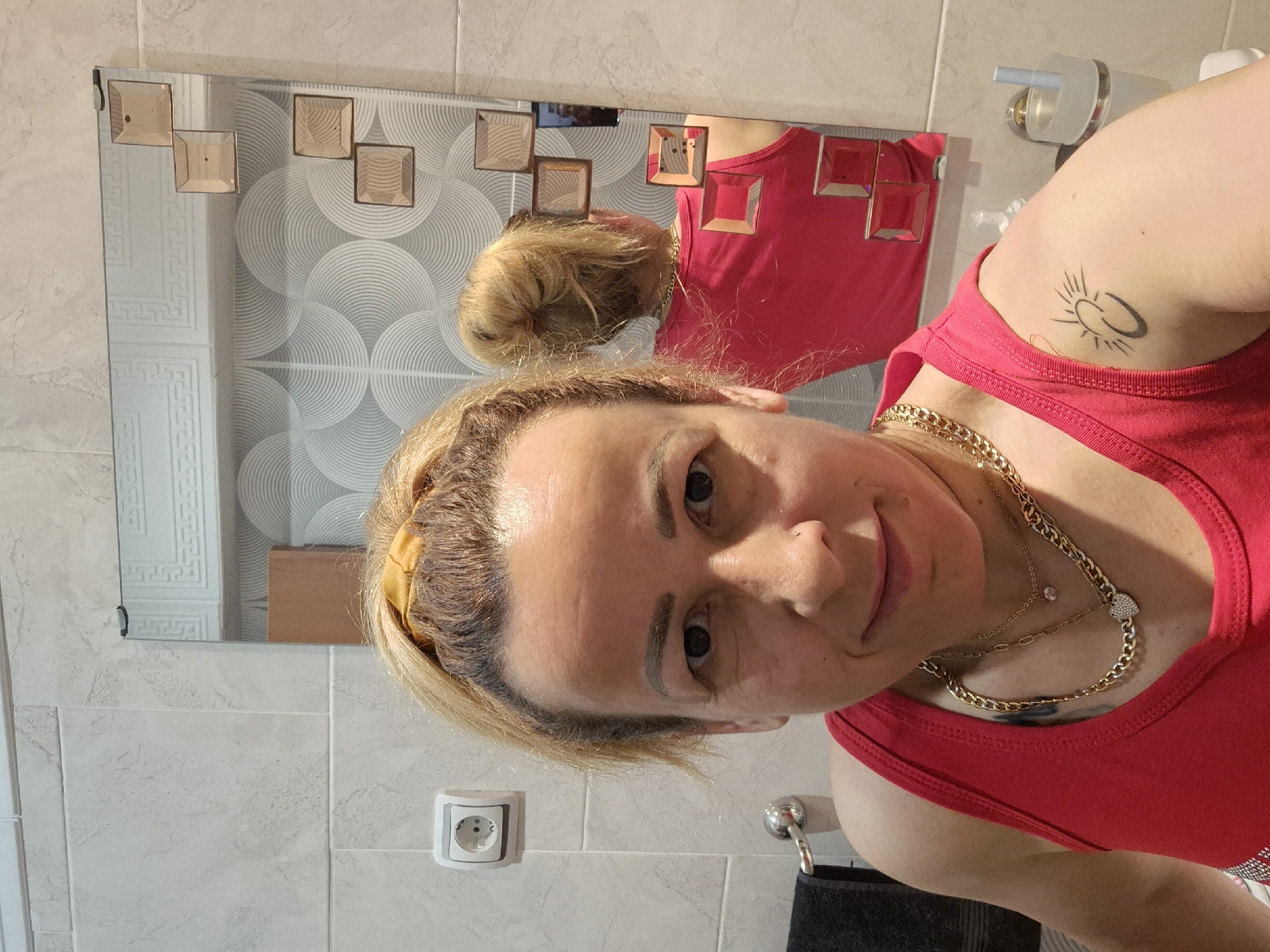 The image captures a selfie taken by a woman in a bathroom, rotated 90 degrees clockwise. The woman, with blonde hair pulled back, wears a red tank top and multiple gold-colored chain necklaces, one featuring a small clear gem and another a heart charm. A tattoo of a sun with a halo is visible on her upper arm or shoulder. She is looking towards the camera with a slight, pleasant smile.
The setting is a bathroom, identifiable by the tiled walls and a mirror. The walls feature light-colored tiles, some plain and some with decorative patterns, including swirling circles and a geometric Greek key-like design. An electrical outlet is visible on the wall. In the background, part of a toilet paper holder with a roll of toilet paper and a metallic towel hook with a dark towel can be seen.
The mirror itself has decorative square accents, possibly amber-colored glass, attached to its surface. The woman's reflection shows the back of her head with hair in a bun and her torso in the red tank top. The bright indoor lighting suggests no specific time of day. No text is visible in the image.