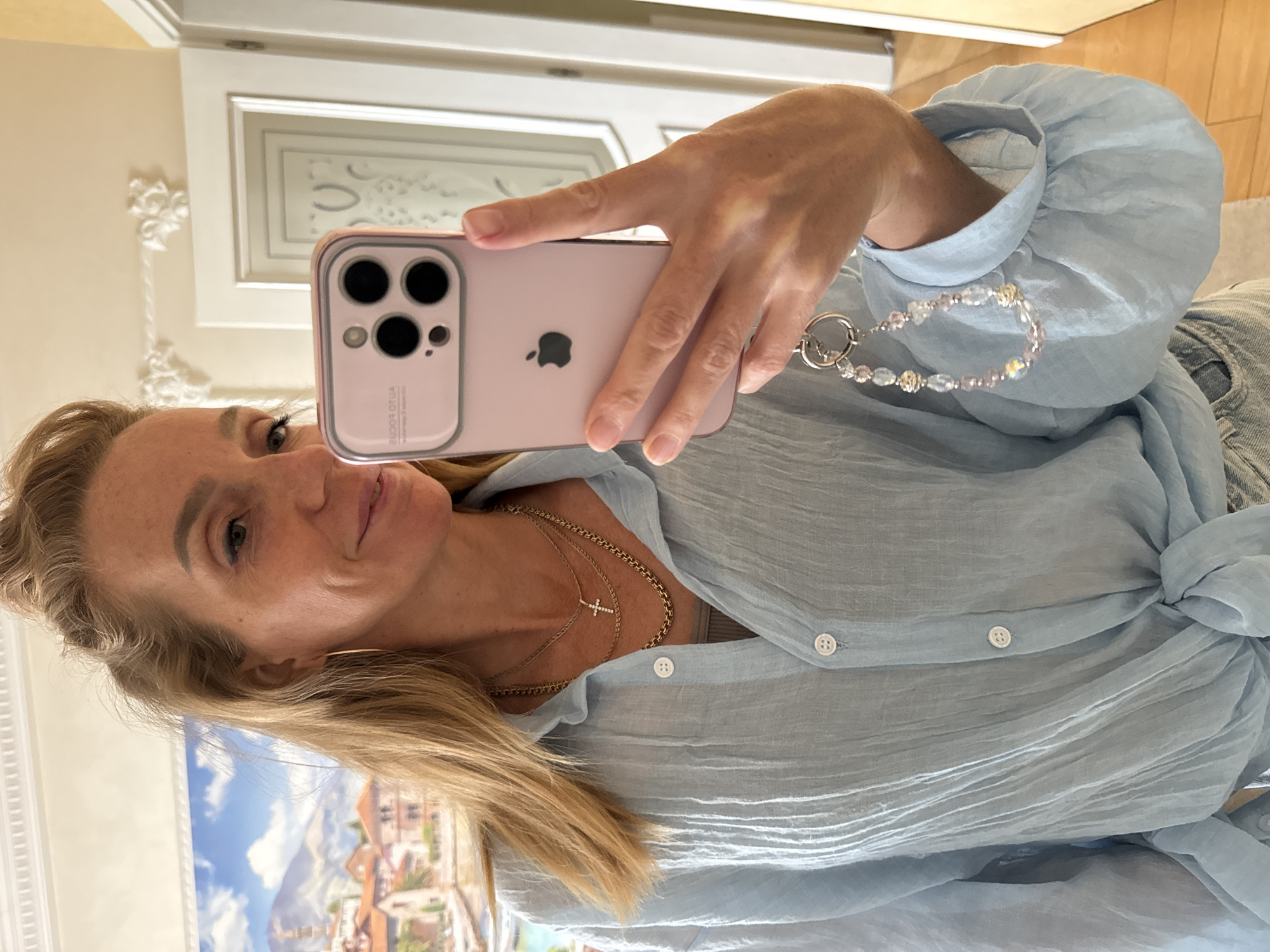 A woman with blonde hair pulled back from her face takes a selfie in a room. She is wearing a light blue, crinkled button-down shirt tied at the waist, a grey tank top underneath, and denim jeans. She has on two gold necklaces, one with a small diamond cross pendant. She is holding a pink iPhone in her right hand, which has a beaded charm attached to its case. The background shows a wall with decorative molding and a closed door. To the left, a painting of a European coastal town with mountains is visible. The lighting suggests it is daytime. The location is not specified as Unknown, Russia.