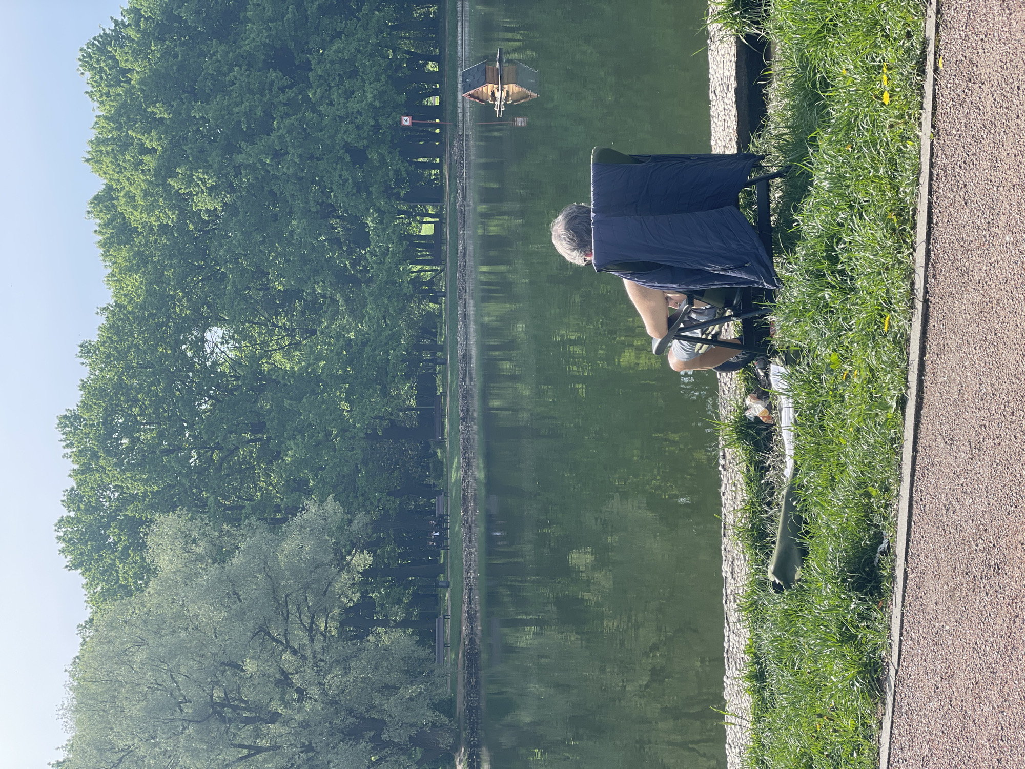 An elderly man sits in a blue folding chair beside a green body of water, possibly a pond or small lake, within a park. The location appears to be in Moscow, Russia, given the presence of trees and a manmade island-like structure with a small roof in the water. The man faces the water with his back to the camera. There's a stone wall separating the water from the grassy bank where the man is sitting, and a gravel walkway is visible beyond the grass. Vegetation includes tall, leafy trees on the far side of the water. There are benches on the far side of the pond. The day appears sunny. A green boot is sitting on the grass.