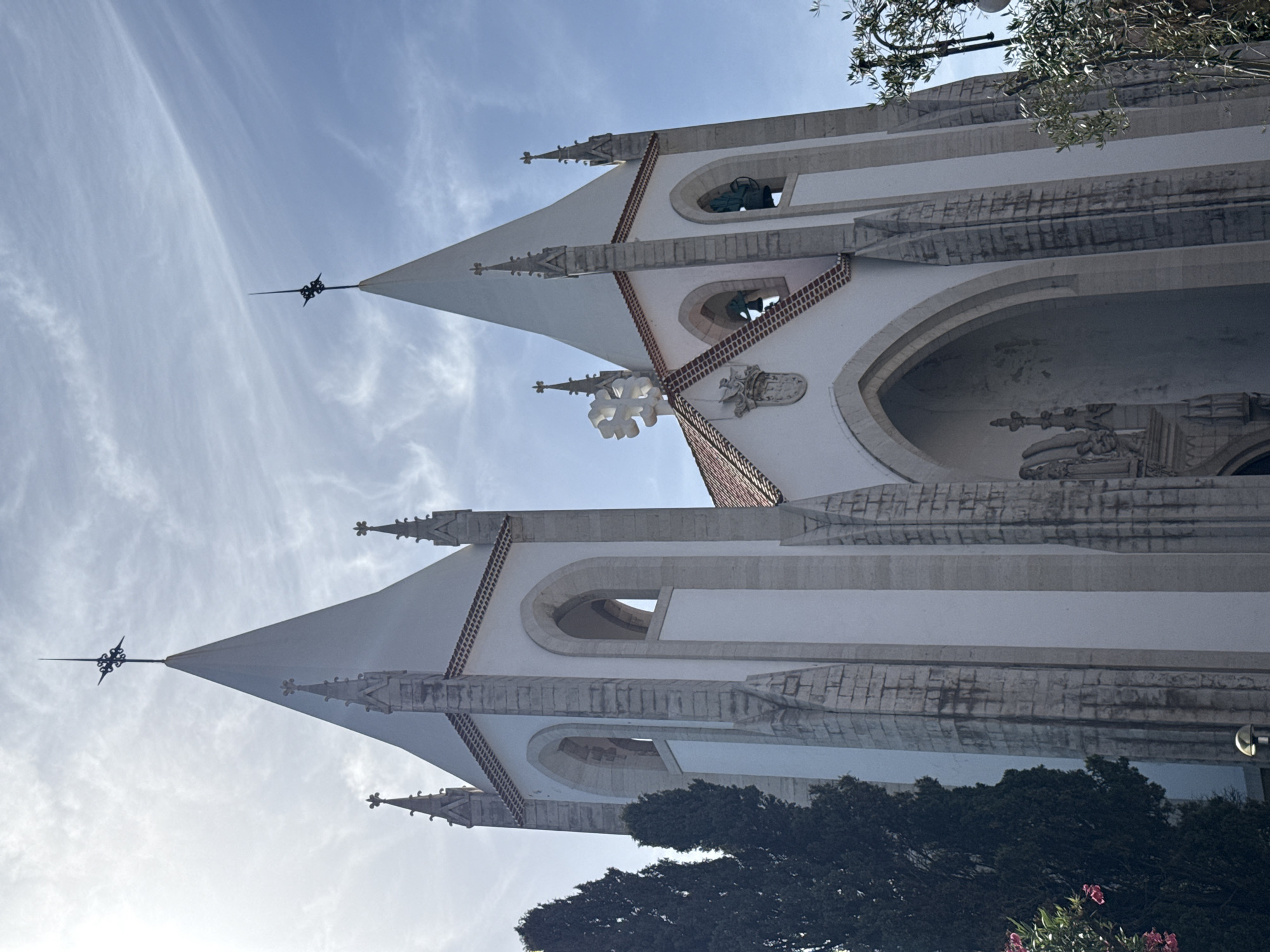 The image showcases the facade of a white building, possibly a church or a grand historical structure, against a bright blue sky with wispy clouds. The architecture features sharp, pointed spires adorned with decorative elements, and arched windows. Several of these windows appear to house bells. The building's design incorporates intricate stone detailing and ornamentation, including what looks like a coat of arms above one of the arched openings. The perspective is from a low angle, looking upwards towards the sky and the upper sections of the building.
In the foreground, partially obscuring the lower part of the building, is a dense cluster of dark green trees. Towards the bottom right corner, a few bright pink flowers are visible, adding a touch of color to the scene. The lighting suggests it is daytime, likely late morning or early afternoon, with the sun illuminating the building from the side. The overall impression is one of serene grandeur and historical significance. The image is oriented vertically. There is no visible text in the image. The location is specified as Lisbon, Portugal.