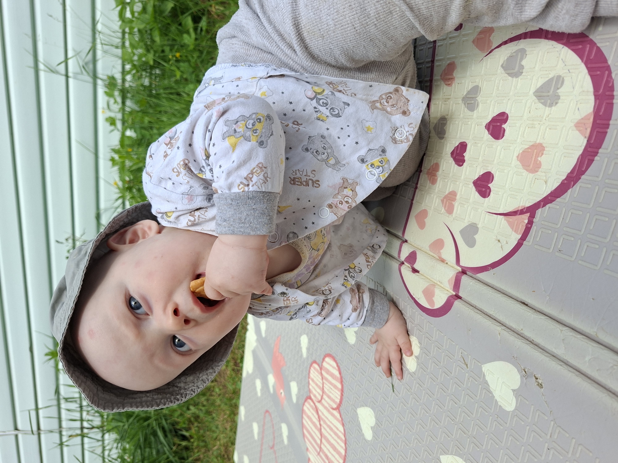 An infant wearing a grey bucket hat, a long-sleeved white shirt with teddy bear graphics, and light grey pants is on its stomach on a grey play mat. The infant is putting a light brown cracker into its mouth. The play mat has heart shapes and cloud patterns. In the background, there is green grass and a light green horizontal striped wall. The overall setting suggests an outdoor environment.