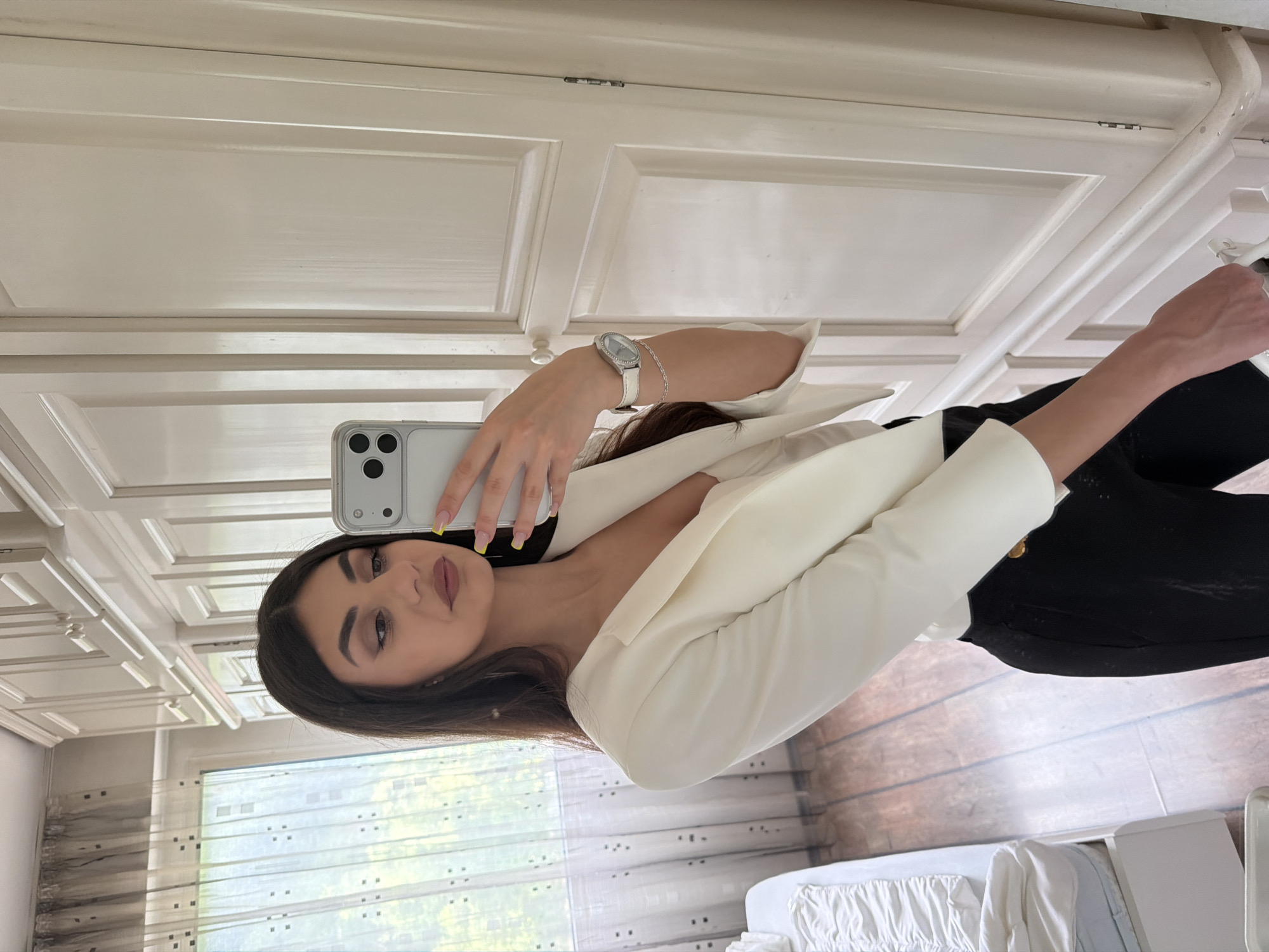 A woman is taking a selfie in front of a mirror. She is wearing a white blazer and black pants. The background includes a white door with panels and a window with sheer curtains. It's impossible to determine if this image was taken in Gabrovo, Bulgaria without additional information or visual cues indicating the location.