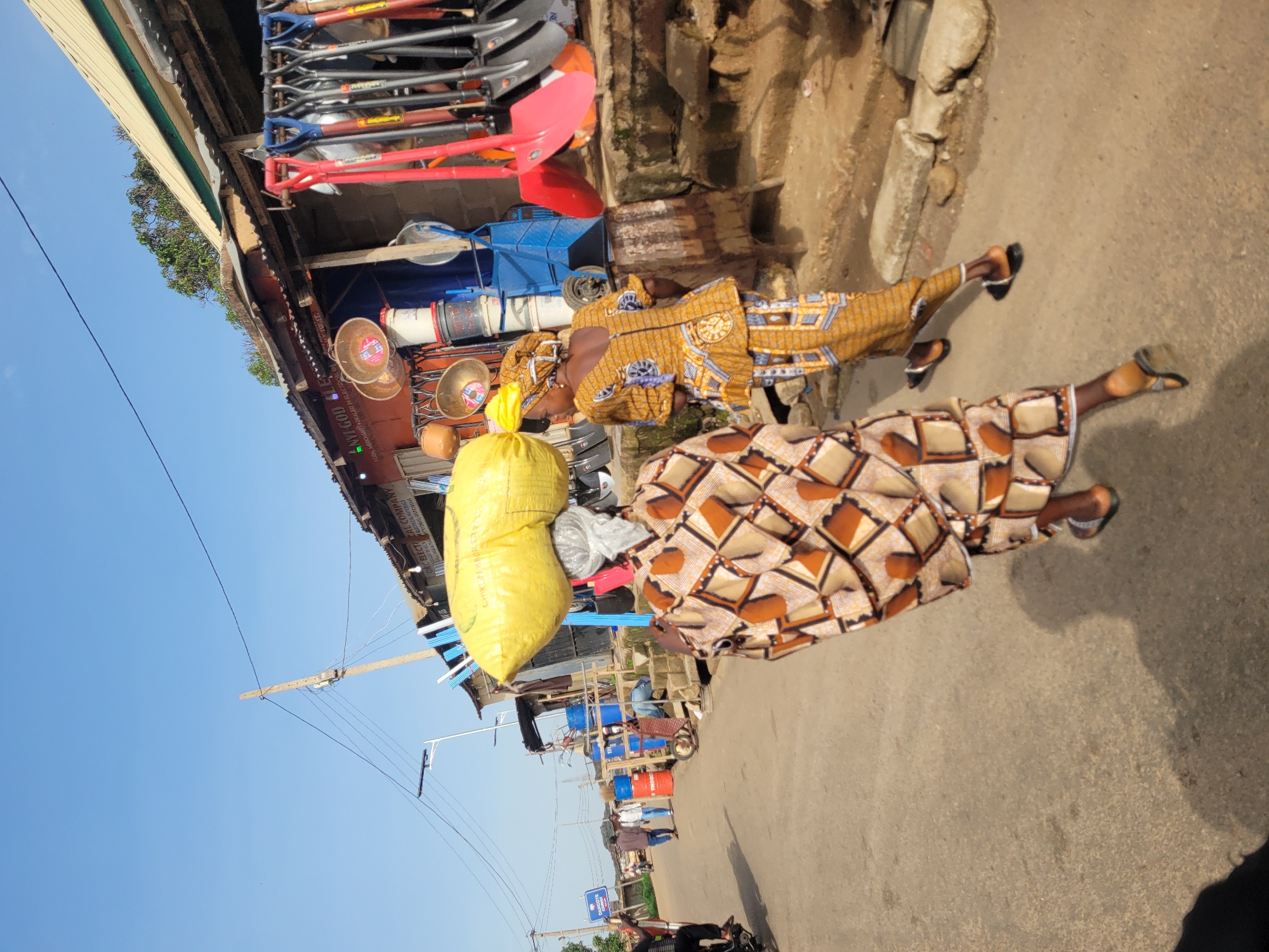 In the city of Gura Top, Nigeria, two women walk down a paved road in front of a market stall. The woman in the foreground, from the back, carries a large yellow bundle balanced on her head, wearing a brown and tan patterned dress and sandals. Beside her, to the right, another woman walks away from the camera, wearing a yellow and blue patterned outfit and sandals. The market stall displays various items, including an assortment of shovels and buckets. The background reveals a clear blue sky and a few trees. The scene depicts everyday life and commerce in a Nigerian urban setting.