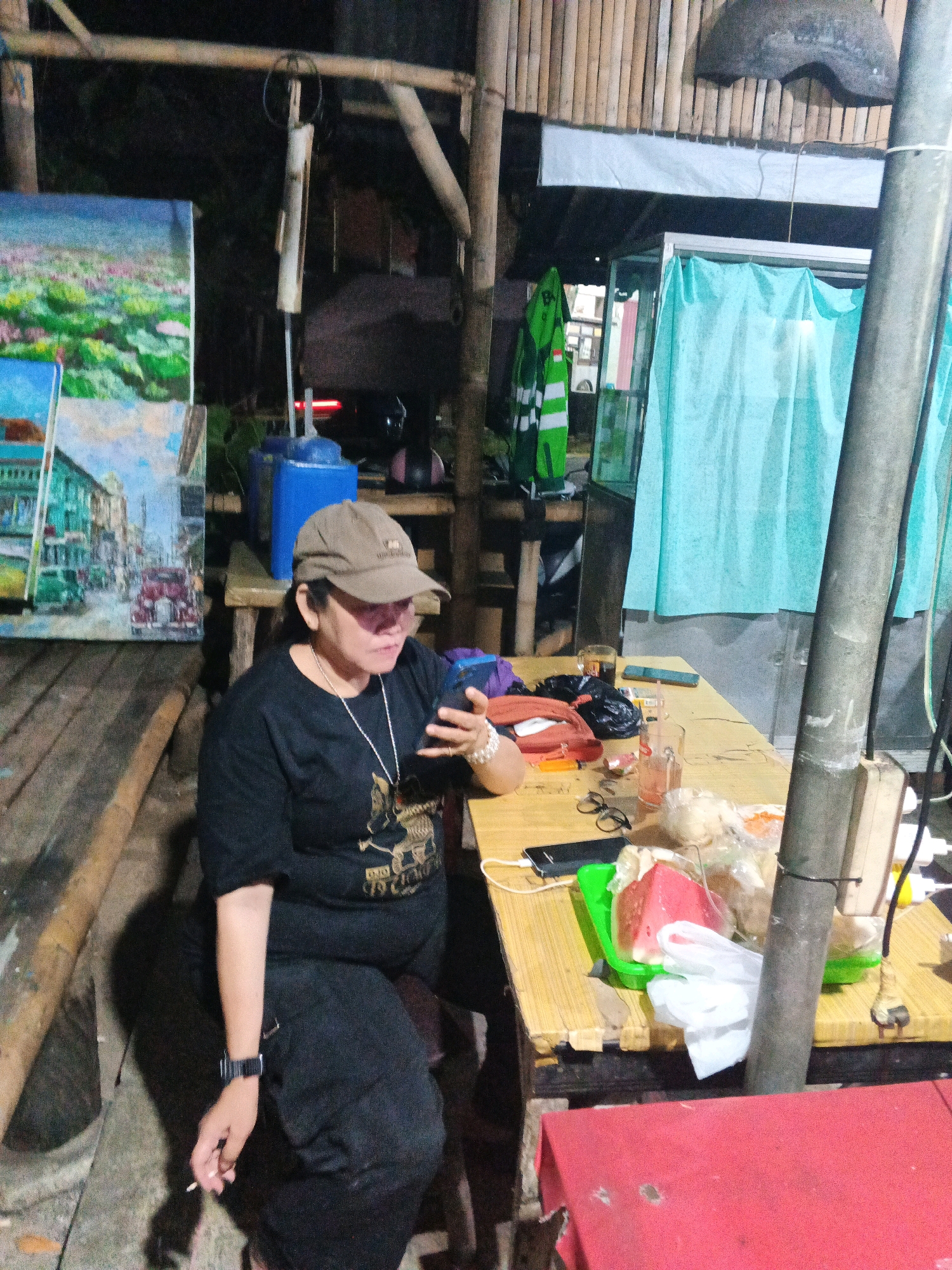 The image depicts a woman seated at a wooden table, appearing to be in an outdoor, possibly informal dining or rest area. She is wearing a black t-shirt, a baseball cap, and dark pants, with a silver necklace. Her right hand is holding a mobile phone, and her left hand is holding a lit cigarette. On the table in front of her are various items including glasses of beverages, food items, a red clutch, a black plastic bag, and a mobile phone. Two paintings are visible to the left of the woman, one depicting a landscape and the other a street scene with vehicles. In the background, there are bamboo structures, a blue water dispenser with a pump, and light blue curtains. The setting appears to be at night or in low light conditions. The presence of elements like the paintings, the outdoor seating, and the bamboo structure suggest a local establishment, potentially a warung or a small eatery, in Ungaran, Indonesia.