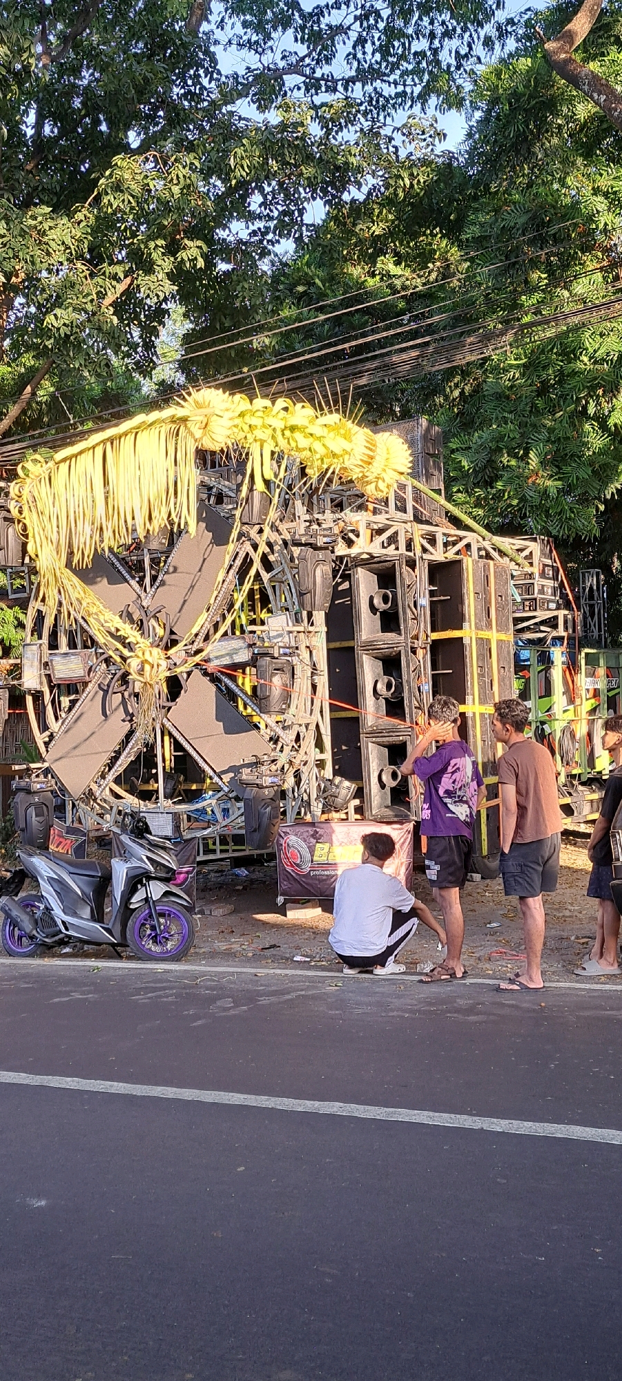 The image depicts a large sound system setup on the side of a road in Prigen, Indonesia. The sound system, adorned with yellow palm fronds, is mounted on a metal frame and includes numerous speakers stacked vertically and horizontally. A silver and purple scooter is parked to the left of the sound system. Four individuals are visible in the scene: one person is squatting in front of a banner, wearing a white t-shirt and black and white patterned pants; another person, wearing a purple t-shirt and black shorts, is standing and touching their head; a third person, in a brown t-shirt and cargo shorts, stands next to them. A fourth person, partially visible on the far right, appears to be wearing dark shorts. The background features lush green trees and utility lines, suggesting an outdoor setting on a sunny day. The ground appears to be asphalt with some debris scattered around.