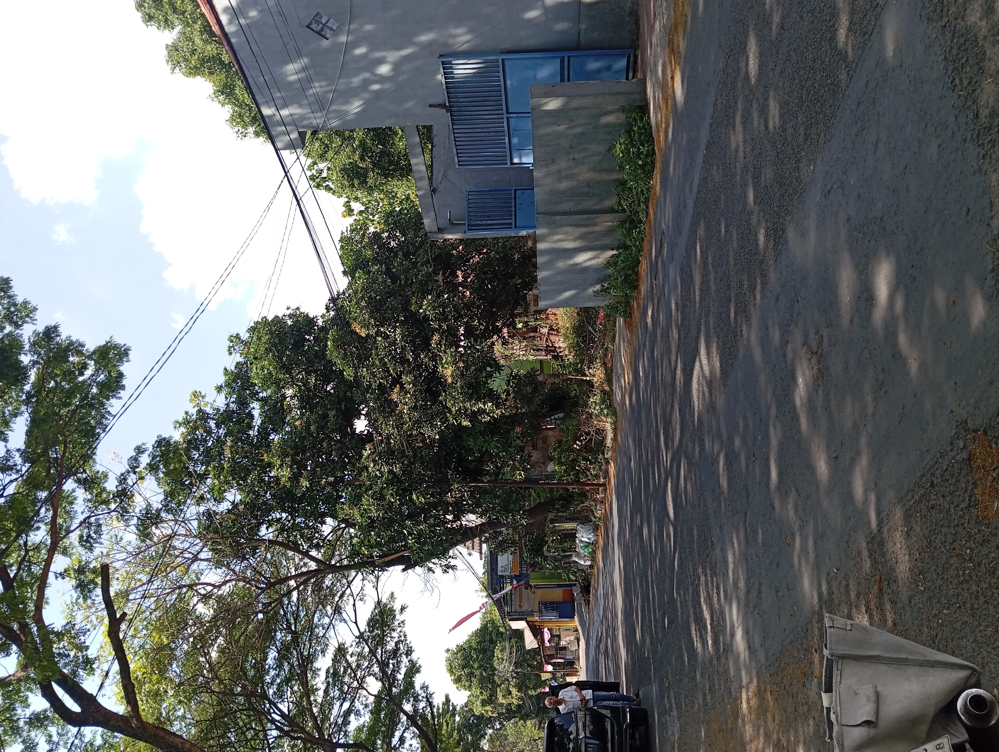 In Karangrejo, Indonesia, a street scene unfolds under a bright, partly cloudy sky. The foreground is dominated by an asphalt road, dappled with sunlight filtering through the canopy of trees lining the left side of the street. A gray, canvas-like object, possibly a large bag or cover, rests on the shoulder of the road in the lower left corner, with a portion of a vehicle's exhaust pipe visible.
Further down the road, a dark-colored SUV is parked on the left, with its rear end towards the viewer. A man in a white shirt and dark pants stands near the rear of the SUV, facing away from the camera. Beyond the SUV, a row of low-rise buildings with colorful awnings and signs line the street. Some of these buildings appear to be shops or small businesses. White sacks are stacked near one of the buildings.
On the right side of the street, a modern-style building with a gray concrete wall and a large blue metal gate is visible. The gate is partially open, revealing a glimpse of the interior. The wall in front of the gate is lower and also made of concrete. Green foliage grows along the base of the wall and the side of the building. Overhead, power lines stretch across the scene, supported by poles that are partially obscured by the lush green trees. The trees themselves are a dominant feature, with large branches and dense foliage creating a canopy that casts shadows on the road. The overall impression is of a quiet, residential or light commercial street in a tropical environment.