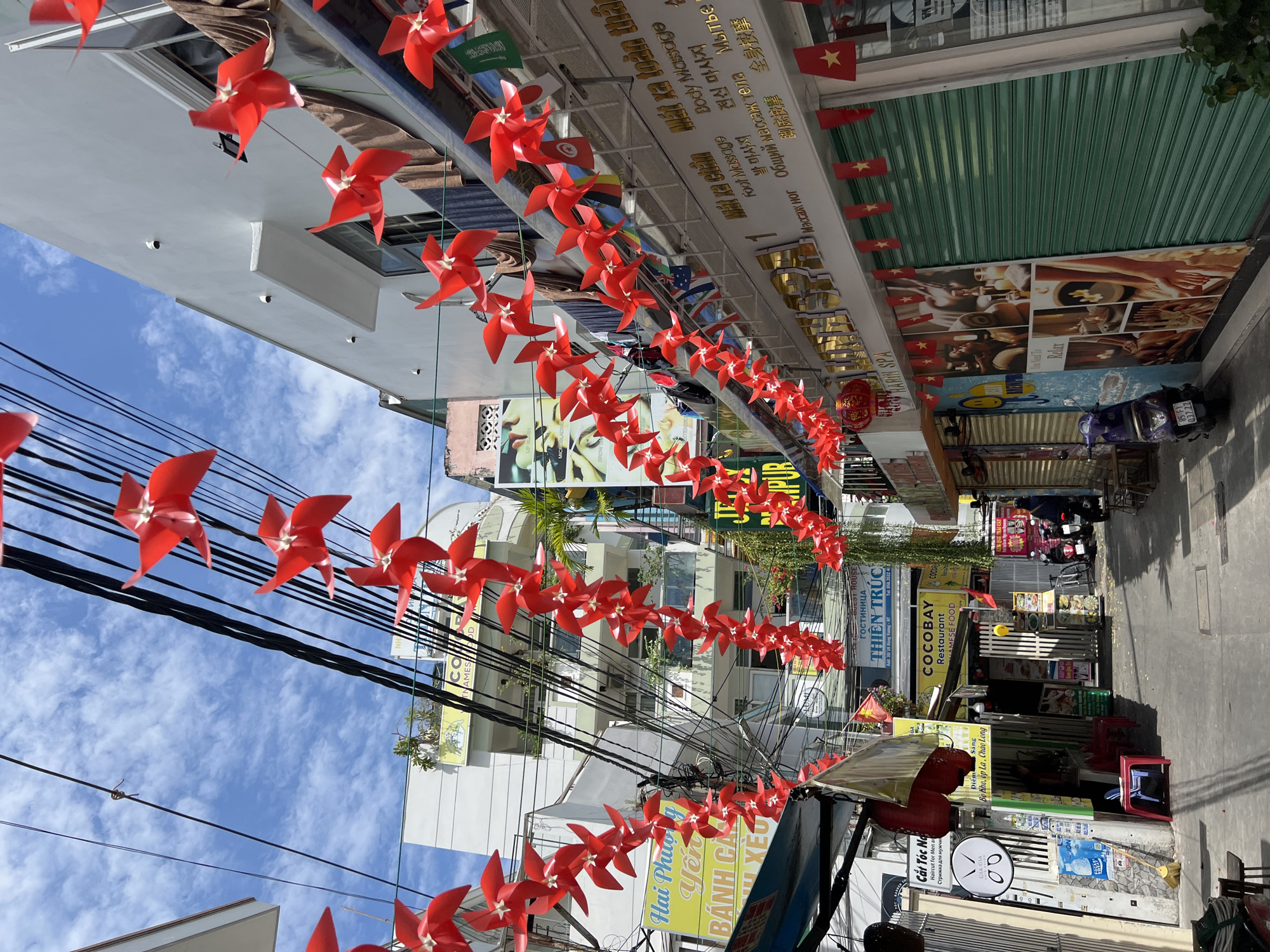 The image captures a narrow street in Nha Trang, Vietnam, decorated with numerous red pinwheels strung across overhead wires. The street is lined with multi-story buildings, many of which house businesses. On the left side, a sign reads "Cắt Tóc Nam Haircut For Men" and "Стрижка для мужчин." Further down, a yellow sign announces "COCOBAY RESTAURANT VIETNAMESE FOOD." On the right side, a business with the prominent text "Mát xa toàn thân Body Massage" and various languages indicating massage services is visible, adorned with small Vietnamese flags. Several green metal shutters cover storefronts, and a purple scooter is parked near one. The ground is paved, and the sky above is partly cloudy. The overall impression is a vibrant, bustling street scene common in Vietnamese cities.