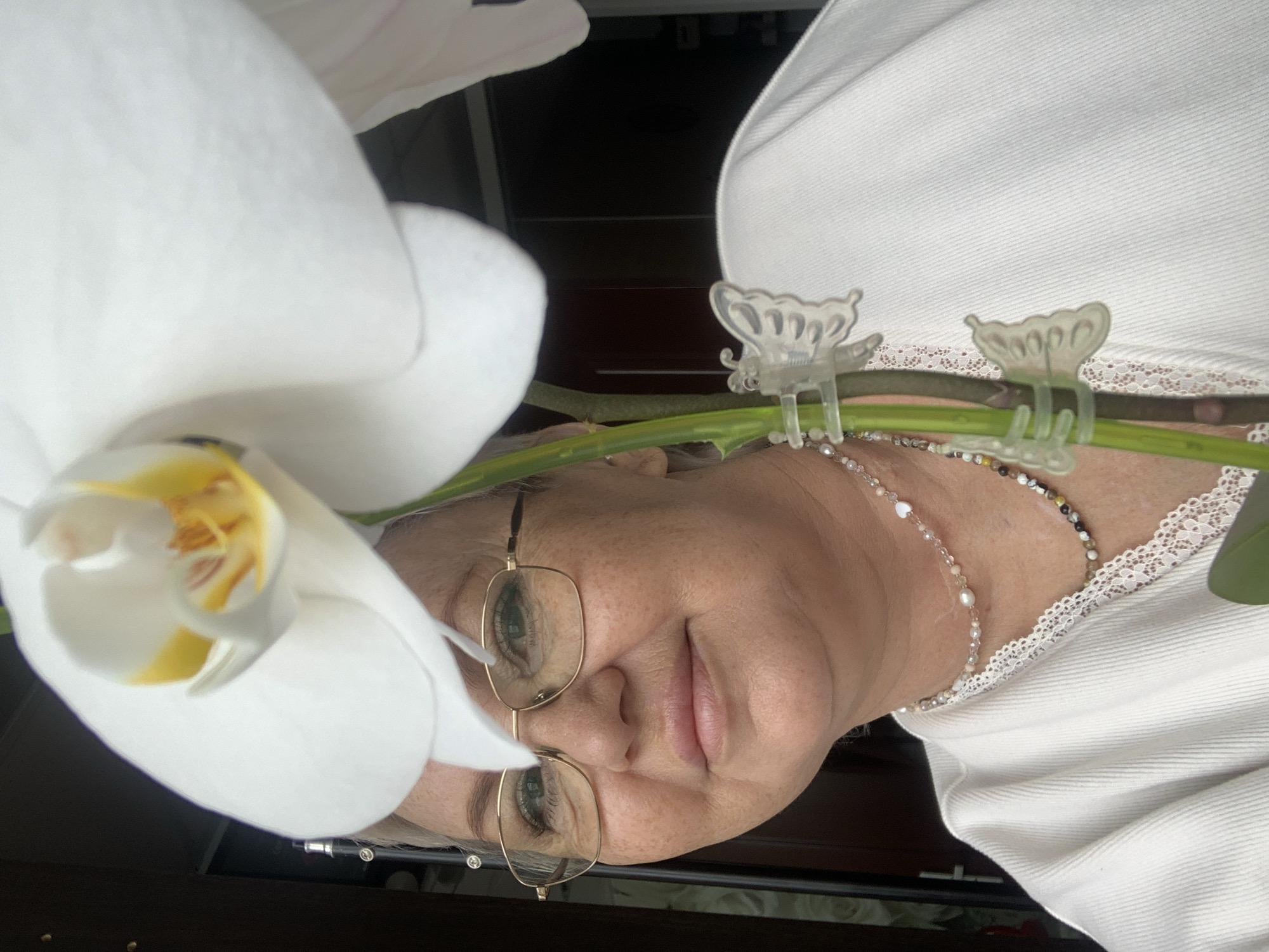 An adult woman with fair skin is photographed indoors, likely in a personal residence, in Chaikovsky, Russia. She has light hair, wears gold-rimmed glasses, and a pearl necklace. She is positioned behind a large white orchid, its petals dominating the foreground. Two small, clear butterfly-shaped clips secure the orchid's green stem. She wears a white garment with lace detailing at the neckline.